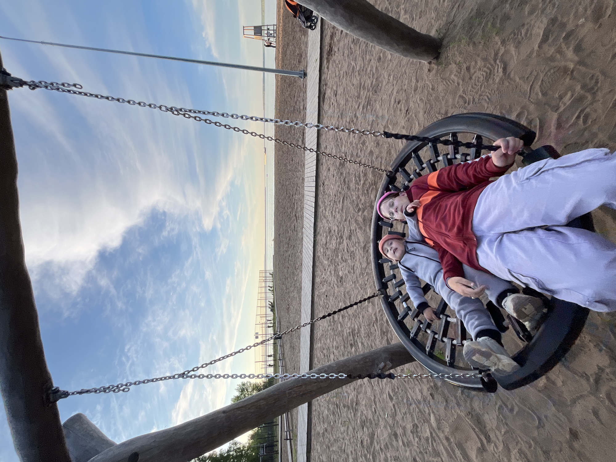 This image captures two children resting in a large, round basket swing on a sandy beach or lakeside playground in Chaikovsky, Russia. The photograph, originally taken with a vertical orientation and rotated 90 degrees counter-clockwise, shows the swing suspended by metal chains from robust wooden support beams.
Two children are visible within the black-framed swing. The child in the foreground wears a red and orange hooded jacket, light grey trousers, and a pink beanie, looking towards the camera with a calm expression. Cuddled beside them is a second, younger child dressed in a light grey tracksuit and a reddish-orange hat, also facing the camera. Both appear relaxed and comfortable in the swing.
The setting features an expanse of sandy ground, suggesting a beach or a playground located near a body of water. A light-colored wooden boardwalk stretches across the sand towards the background. In the distance, a wide body of water, likely the Votkinsk Reservoir, meets the horizon under a spacious sky. The sky is largely clear, with soft, wispy clouds. A prominent orange A-frame structure, possibly a lifeguard stand, is visible on the sandy shore beyond the boardwalk. The soft, warm illumination suggests the photograph was taken during late afternoon or early evening. No text is visible in the image.