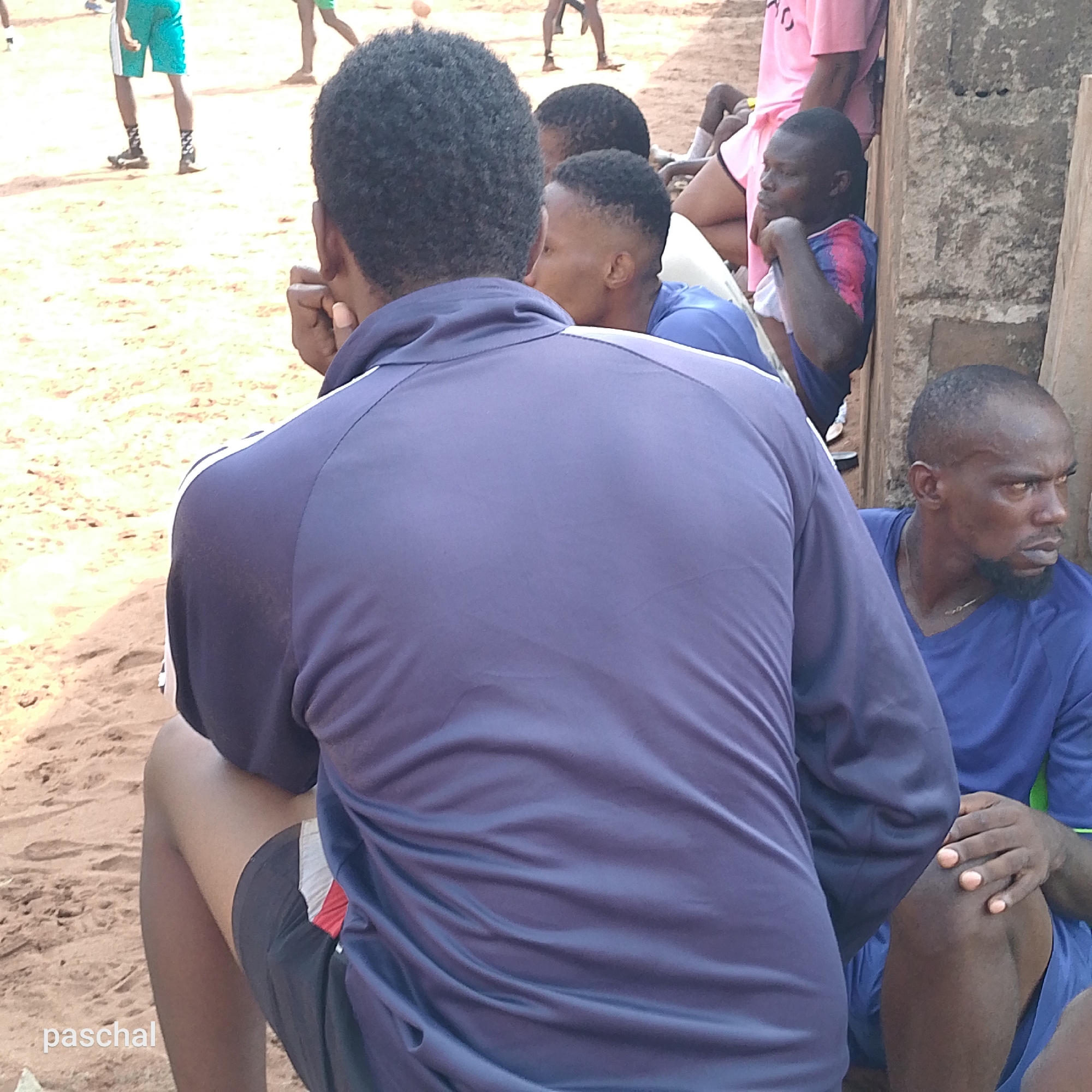 In Ukwu-Abwa, Nigeria, several people are sitting in an outdoor area with a sandy ground. Some of them are wearing sports attire, with a few men in blue jerseys. The individuals are likely watching something happening off-camera. The scene includes part of a concrete wall or structure. Some men are sitting resting, others are standing in the background.