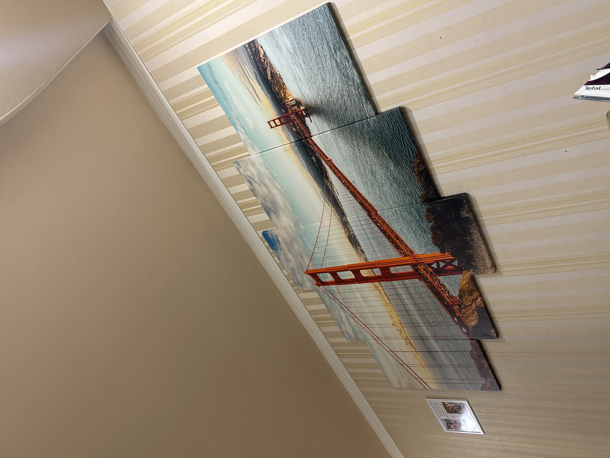 A portrait-oriented shot shows a wall with a print of the Golden Gate Bridge. The wall has a striped wallpaper pattern in alternating light and dark beige. The print is composed of five square canvases arranged in a staggered pattern. The bridge is red and extends diagonally across the canvases. The water surrounding the bridge is a light blue. The wall on the left is a solid, light beige color. On the right side of the wall, near the top, is a corner of what appears to be a box with the word "Tefal" visible. Near the bottom, there is a small, framed picture on the wall, but the details are difficult to discern. The photo is taken from a slightly angled perspective, and the location is not identifiable as Yerevan, Armenia based on the visual information provided.