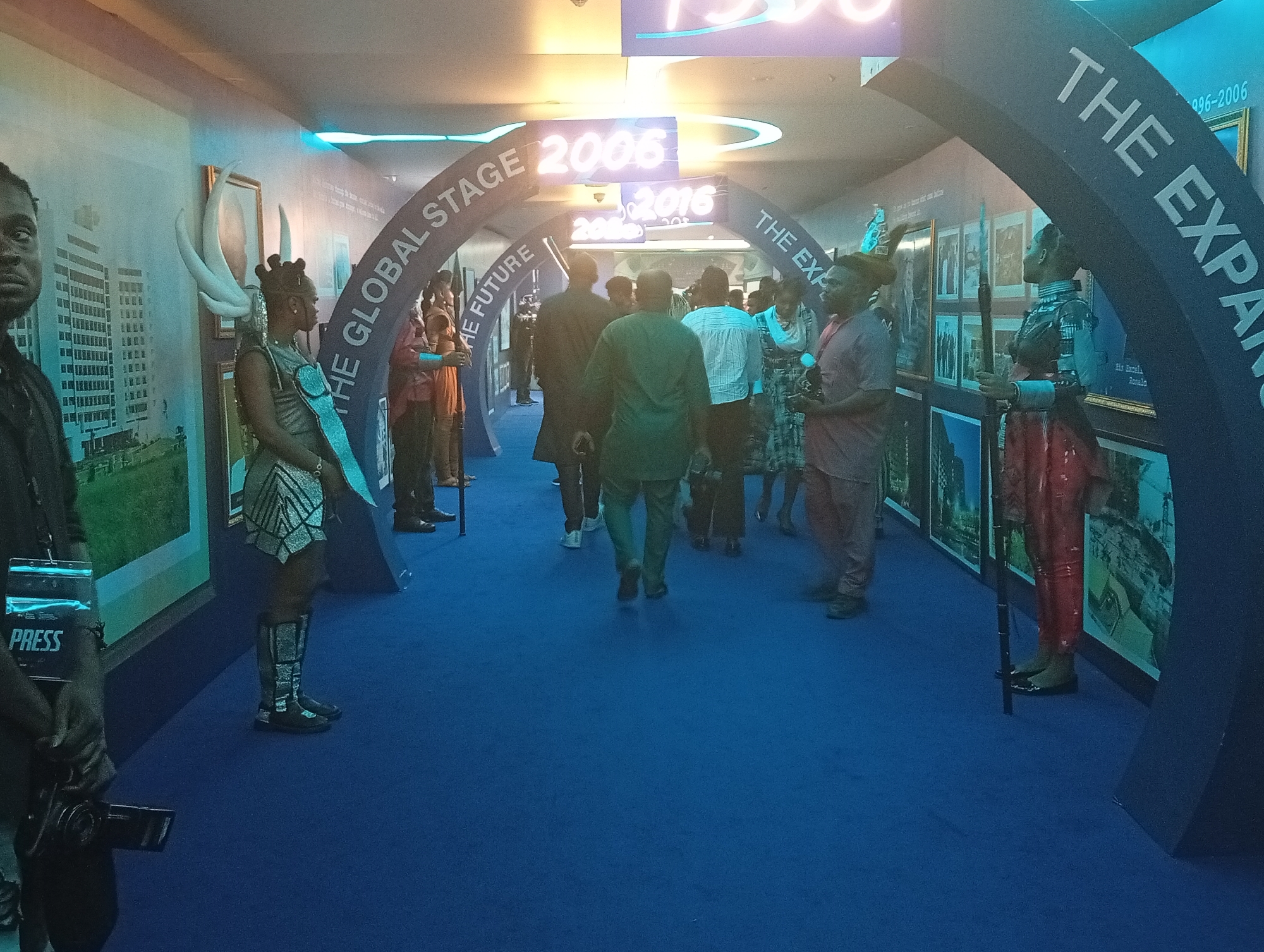 Here is a forensic description of the image:
**Scene:** The image appears to be taken indoors at an event or exhibition, as suggested by the carpeted floor, arched entryways with text, and framed images lining the walls. The lighting is a combination of natural and artificial, with some elements backlit.
**People:** Several people are present, including some walking through the arched entryway and others standing to the side. Of note, there is a person in a costume with elaborate headwear and attire to the left, another person in costume with a spear stands to the right. A person is holding a press pass with a camera to the left.
**Objects:** Arched entryways with text such as "The Global Stage" and "The Expansion" are visible. Dates like "2006" and "2016" are displayed above one archway. Framed images, likely photographs or artwork, are hung on the walls.
**Actions:** Some people are walking through the archways, suggesting they are exploring the exhibition or event. The people in costumes are holding spears standing guard on either side of the corridor.
**Interactions:** People are engaged in walking through the arched entries and interacting with the displays on the walls.
**Environment:** The setting seems to be within a building or exhibition hall. It appears to be an open area, possibly a corridor or passage. The overall atmosphere suggests a cultural event or exhibition. The OCR notes "His Exceli Ronala." It may be the exhibition is celebrating this person.
**Location Clues:** There is a clue that this scene may be taking place in Itirin, Nigeria. This clue is not visually available in the image, and is based on the user's request.