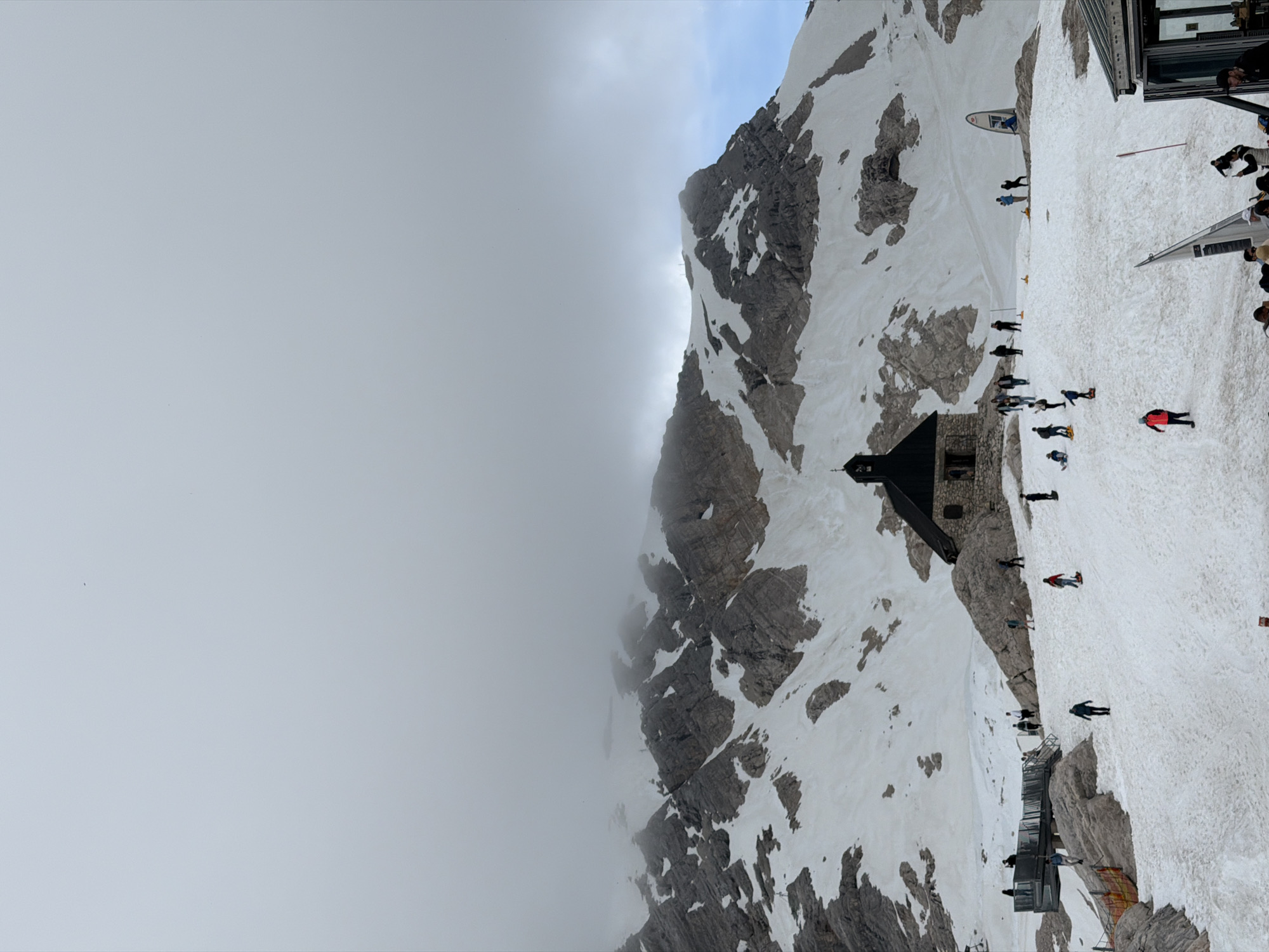 The image depicts a view of the Zugspitze, Germany's highest peak, located near Garmisch-Partenkirchen. The scene is set on a snowy mountain slope, with a small stone building resembling a chapel at the center. Several individuals are scattered across the snow-covered terrain, some appearing to be walking, while others might be engaged in recreational activities. The background features rugged, snow-capped mountains partially obscured by clouds, indicating a high-altitude environment. A viewing platform or walkway is visible to the left, with a few people on it. The overall atmosphere suggests a tourist destination, likely popular for its mountain scenery and outdoor activities.