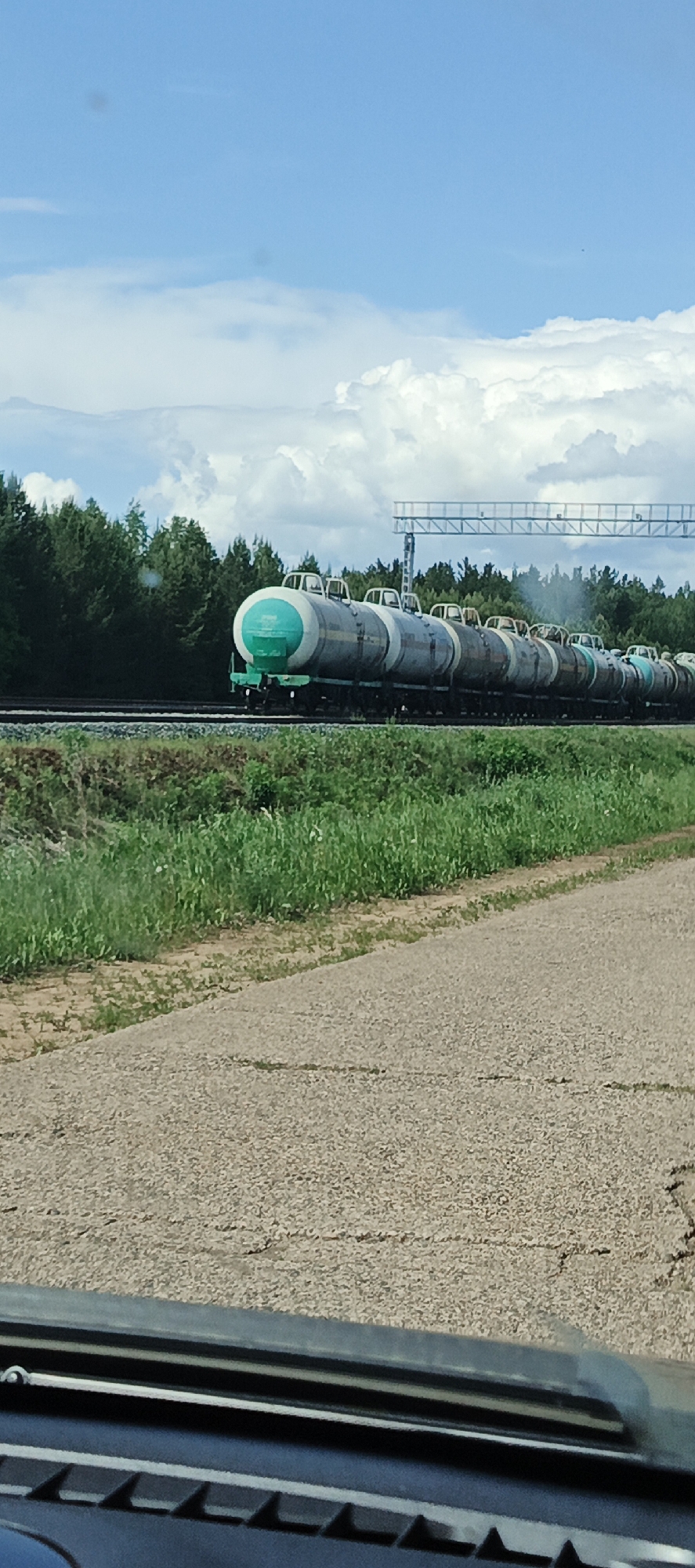 The image depicts a train composed of multiple tanker cars passing through Kauchuk Stantsiya, Russia. The train is positioned on a railway track that runs parallel to a paved road. The tanker cars are light gray with a rounded cylindrical shape, and the lead car has a distinctive bright green circular end. Dense green vegetation, including trees and grass, lines the background and separates the railway from the blue sky with scattered white clouds. The image is taken from inside a vehicle, with part of the dashboard visible at the bottom of the frame.