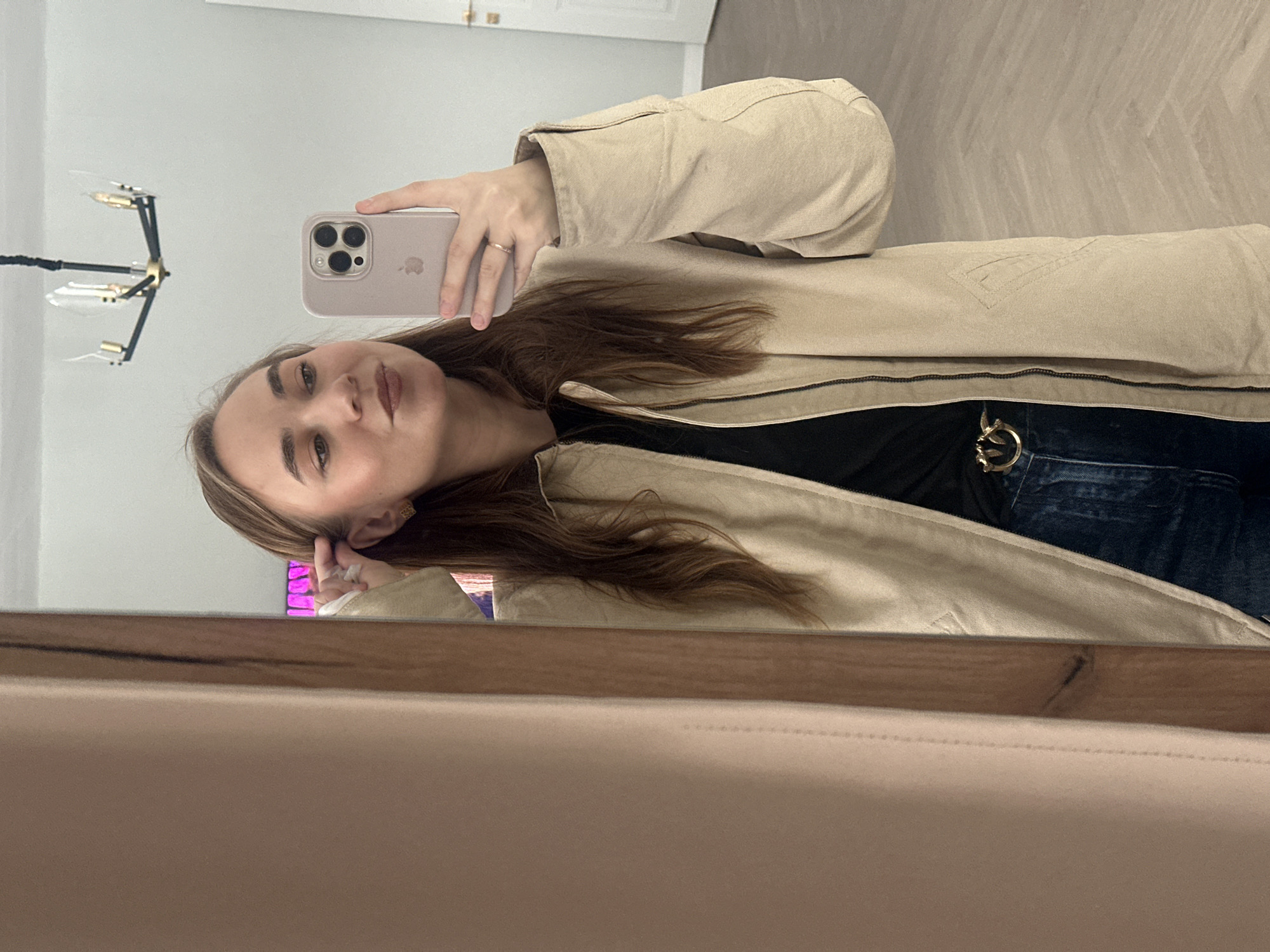 A young woman with light brown hair is taking a selfie in front of a mirror. She's wearing a tan jacket over a black top and jeans with a gold belt buckle. She is holding a modern smartphone with multiple camera lenses. A light fixture with a modern design hangs from the ceiling in the background. There is no information visible that connects the image to Kharnavy, Russia. The background includes a white door and light beige flooring. The mirror's frame is visible at the bottom of the image, constructed from a light-colored wood.