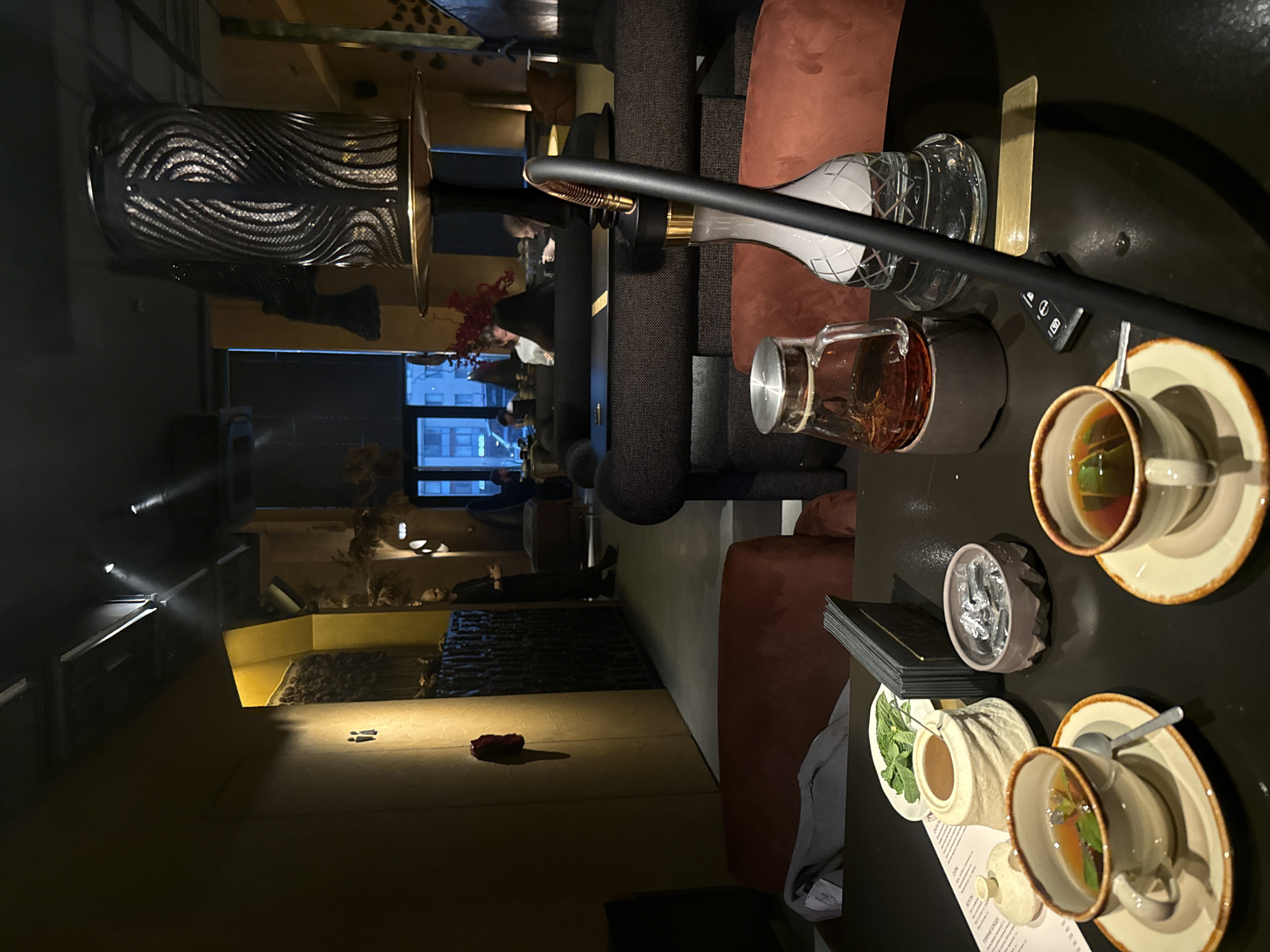 This is an indoor scene, possibly within a restaurant or lounge. The image shows a hookah setup, with the base positioned towards the upper left and the hose extending across the frame. A table setting in the foreground includes teacups on saucers, a teapot, a small dish containing foil, a pot of mint leaves, and a menu. The furniture consists of plush seating, including armchairs and upholstered benches. In the background, through the window, a building in Khimki, Russia, can be seen. Several patrons are seated further inside, and some decorative items are also present.