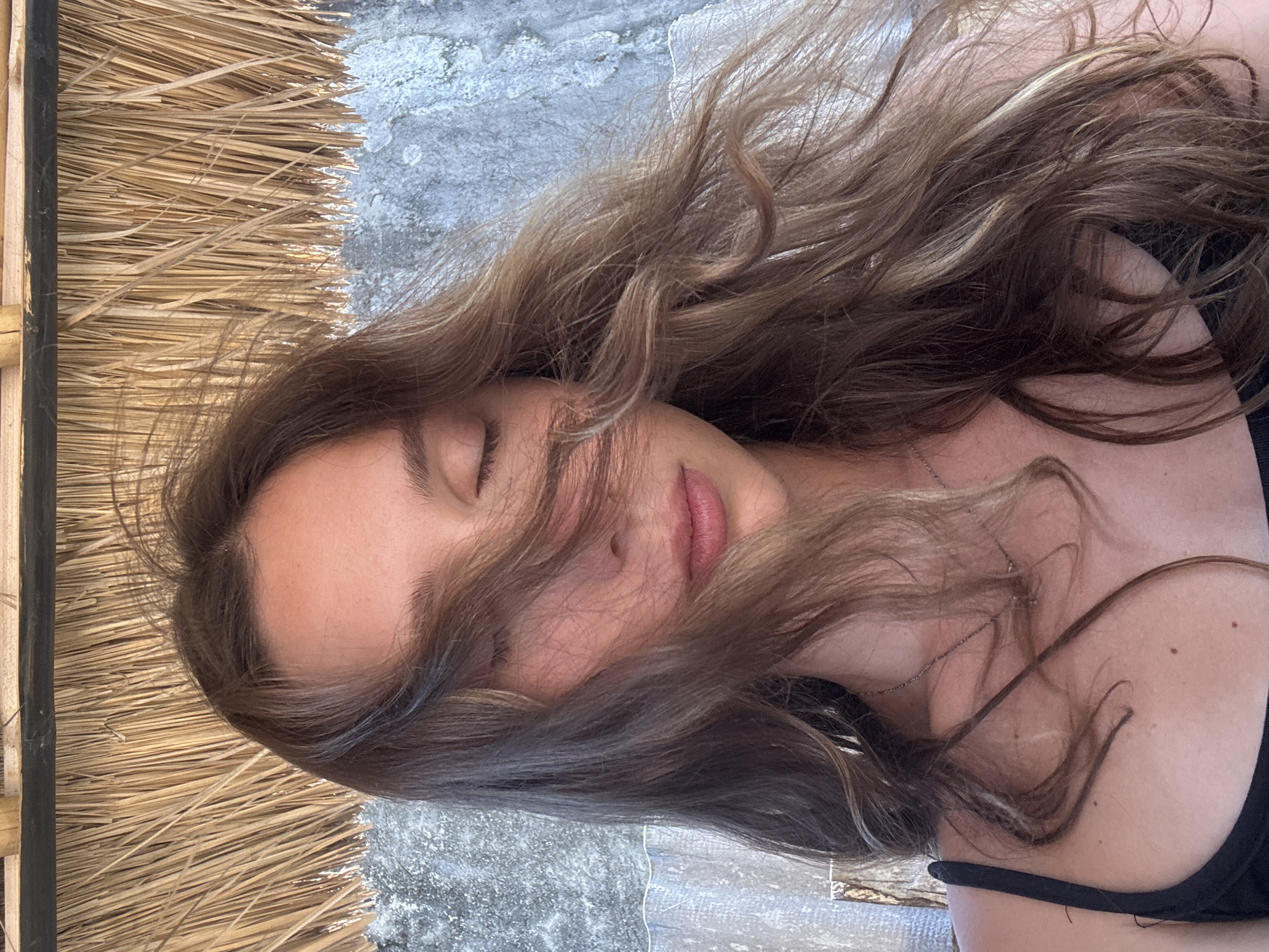 The image shows a close-up of a young woman from the chest up, taken outdoors. She is wearing a black tank top and a silver necklace. Her eyes are closed, and her long, brown hair is blown across her face by the wind. Behind her, there is a textured grey wall and a thatched roof structure. The lighting suggests it is daytime. The location is not specified in the image.