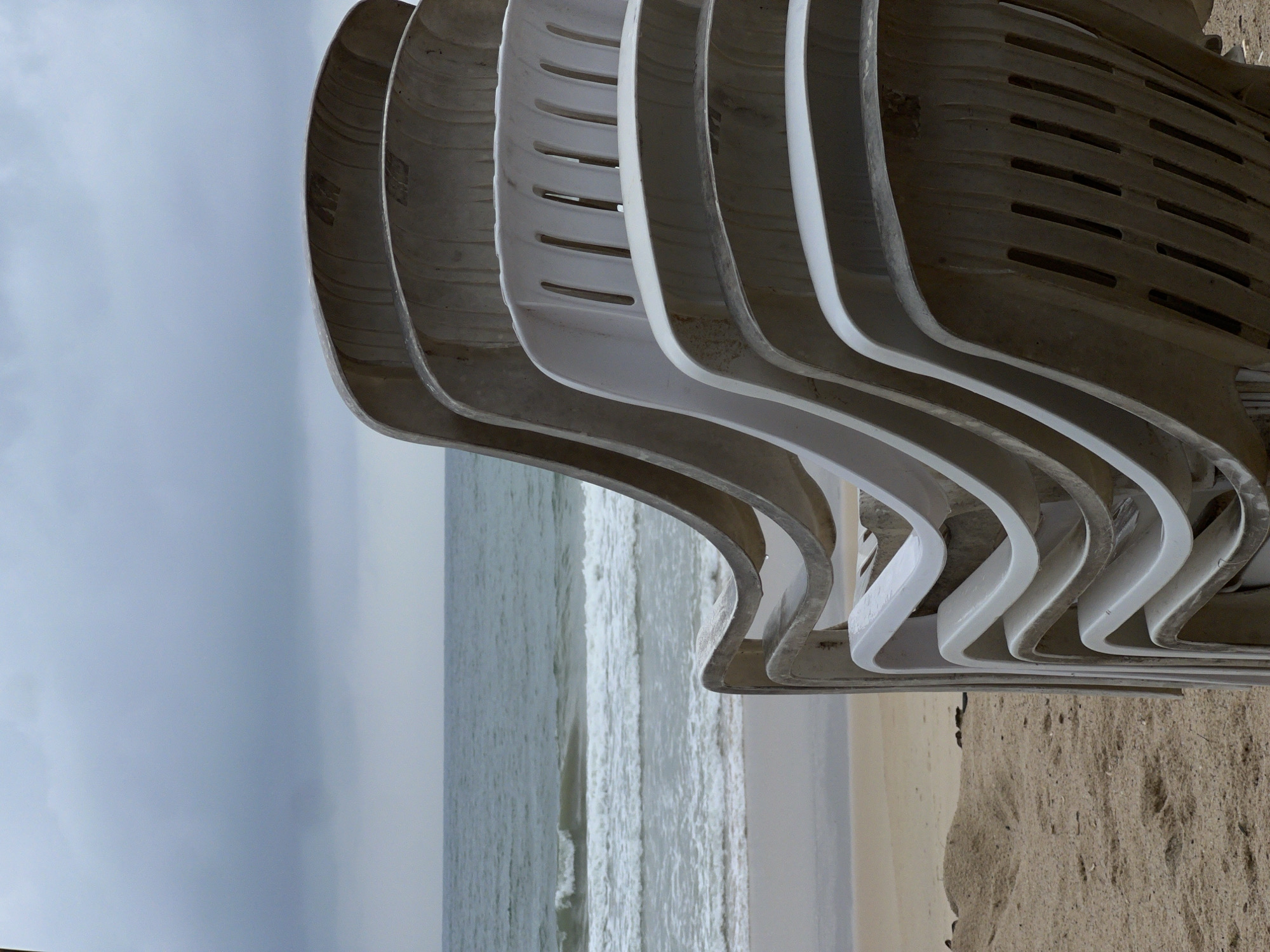 The image captures a coastal scene, likely a beach, under an overcast sky. In the foreground, a stack of white plastic chairs dominates the view. The chairs are stacked neatly, with their curved backs creating a series of repeating lines and negative spaces. Behind the chairs, the scene opens up to reveal the ocean, its surface slightly choppy. The sandy beach is visible in the lower right corner, meeting the water's edge. The image lacks any discernible human presence, emphasizing the stillness and potential solitude of the scene.