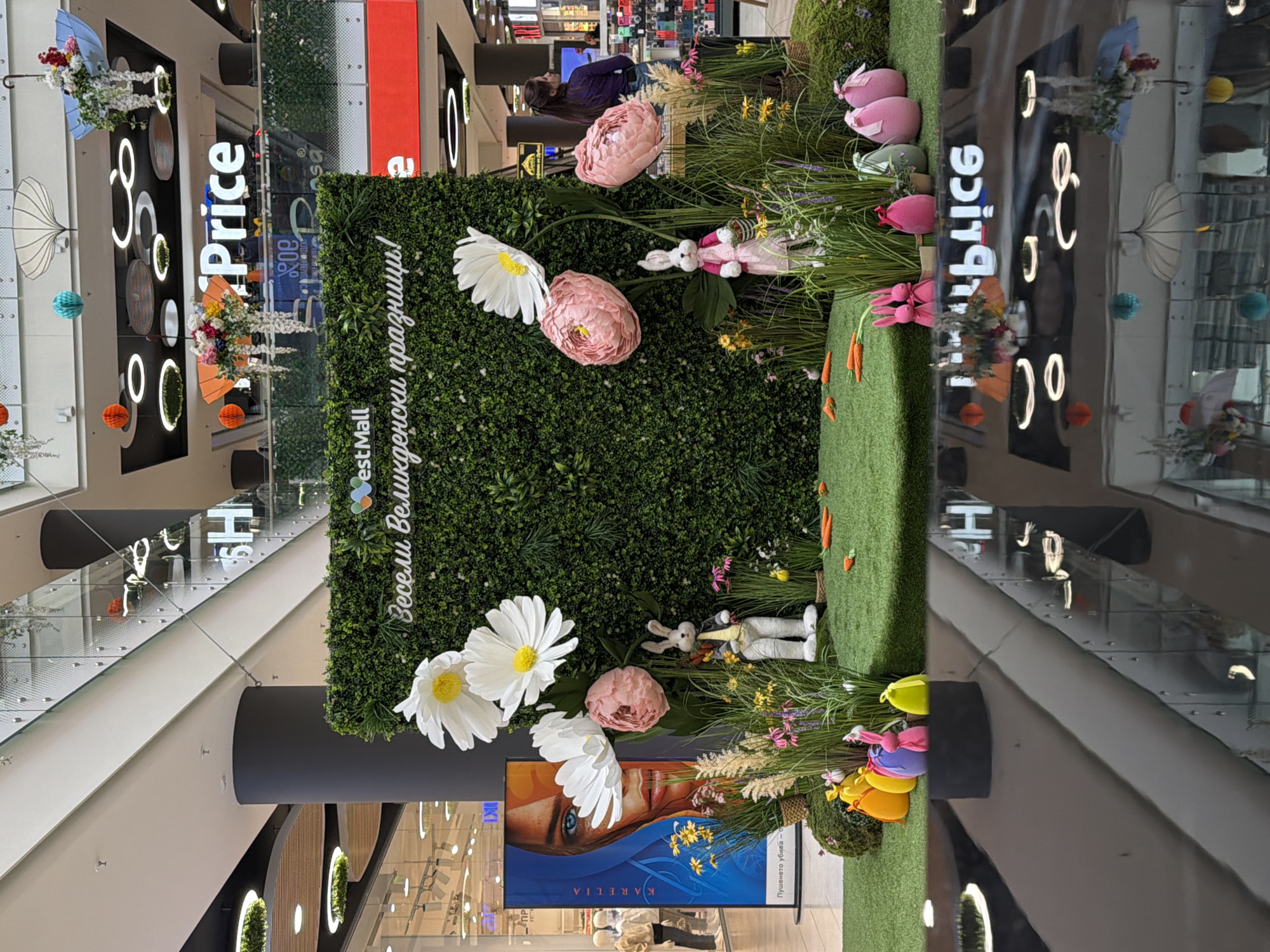 Here is a detailed description of the image:
The image shows an indoor scene, likely a shopping mall in an unidentified location in Unknown, Unknown. The area is decorated for an event, possibly Easter, given the presence of Easter-themed decorations.
The decor includes a large green wall covered in artificial grass and foliage. On this wall, there are various artificial flowers, including large daisies and pink peonies. Easter-themed decorations, like bunnies and eggs, are arranged among the flowers and grass.
On the mall's side, there are signs indicating "Price," which may suggest a store or price comparison service. There are also additional mall-related markings.
A large advertisement or screen displays a woman's face in blue tones. This advertising screen is part of the Easter setup.
People can be seen walking around in the background, indicating that the scene is a public space.