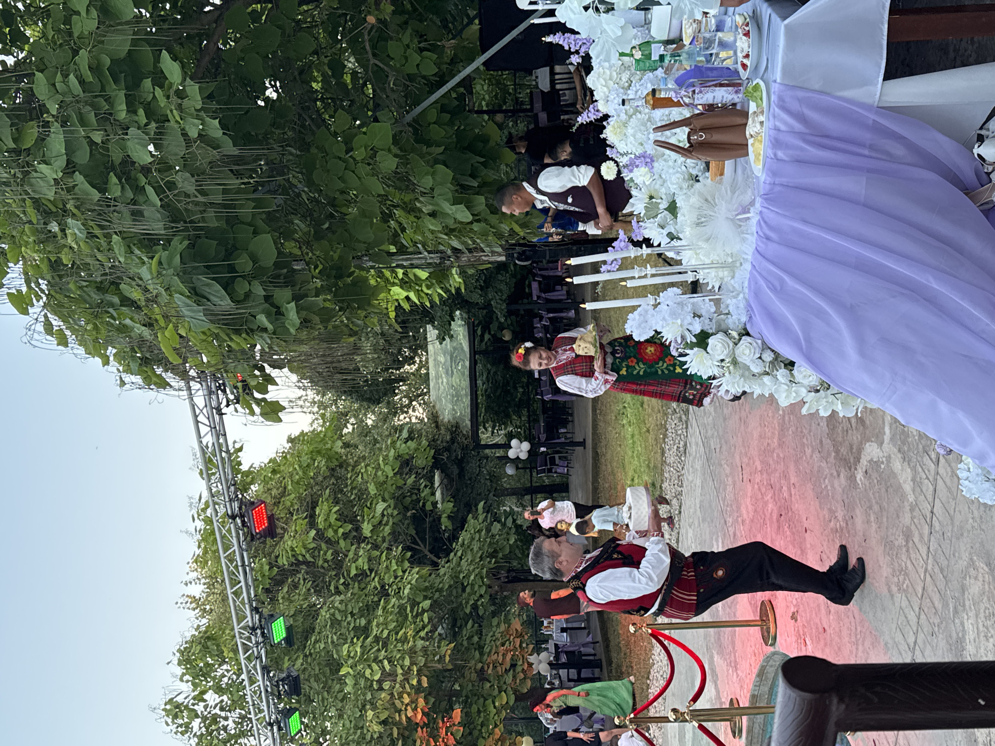 In Sredets, Bulgaria, an outdoor event is underway, possibly a wedding reception, set against a backdrop of lush greenery and a clear sky. A man in traditional Bulgarian attire, a red vest with intricate embroidery over a white shirt and black pants, carries a cake. To his right, a woman in a similar traditional outfit, featuring a red and white plaid vest over a white blouse and a patterned skirt, holds a tray with what appears to be food. In the background, several tables are set up with white tablecloths and chairs, some adorned with white and purple floral arrangements and balloons. Lighting rigs are visible overhead, suggesting the event continues into the evening. The ground is paved with stone and gravel.