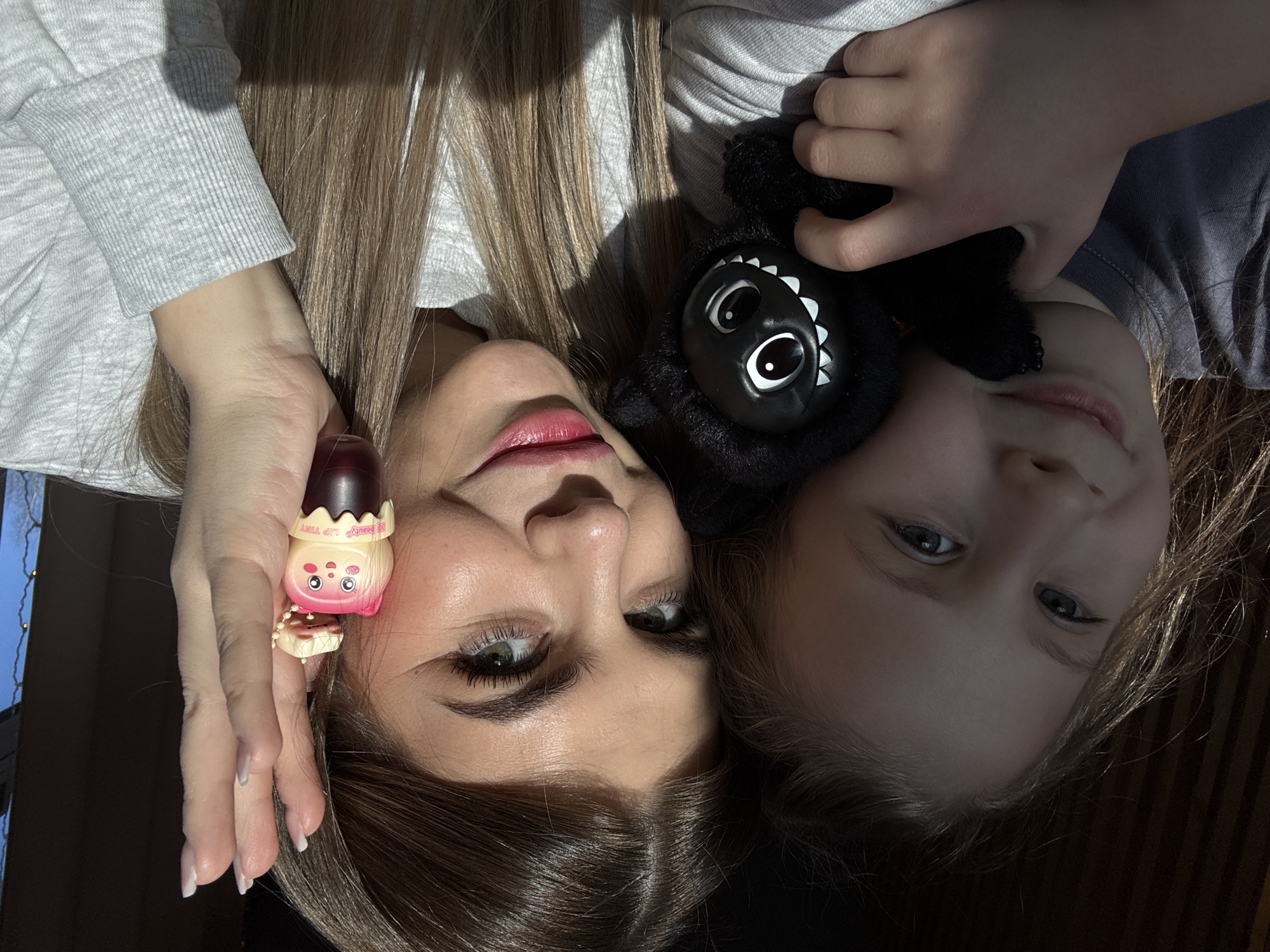This image depicts two females, an adult woman and a young girl, in what appears to be an indoor setting in Chaikovsky, Russia. The adult woman, with light brown hair and a light grey long-sleeved top, is positioned on the right, looking directly at the camera with a smile. She holds a pink and red "BB BEAUTY LIP TINT" container, shaped like a cartoon character, in her right hand near her right ear. Her fingernails have a white tip manicure. The young girl, with blonde hair and a dark grey or blue t-shirt, is on the left, also smiling and looking at the camera. She cradles a black plush toy with prominent white eyes and teeth against her chest with both hands. Both individuals are illuminated by strong, natural sunlight coming from the right. The background is mostly dark and indistinct, with vertical elements on the left and a glimpse of a blue sky and what may be tree branches or faint outdoor lights visible through an opening on the far right.