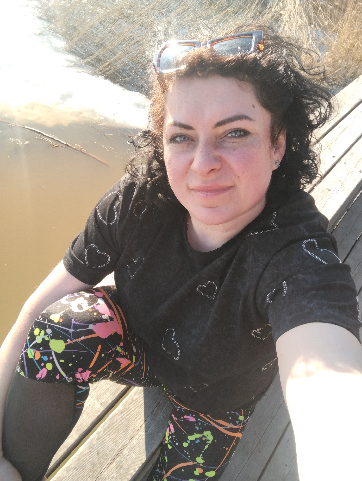 A selfie taken on a sunny day shows a woman with curly dark hair and sunglasses perched on her head, sitting on a wooden platform. She wears a black t-shirt with heart designs and black leggings with a paint splatter pattern underneath. Behind her is a body of water and some vegetation. The exact location is within the city of Votkinsk, Russia, but the immediate setting appears to be a lakeside or riverside wooden structure.