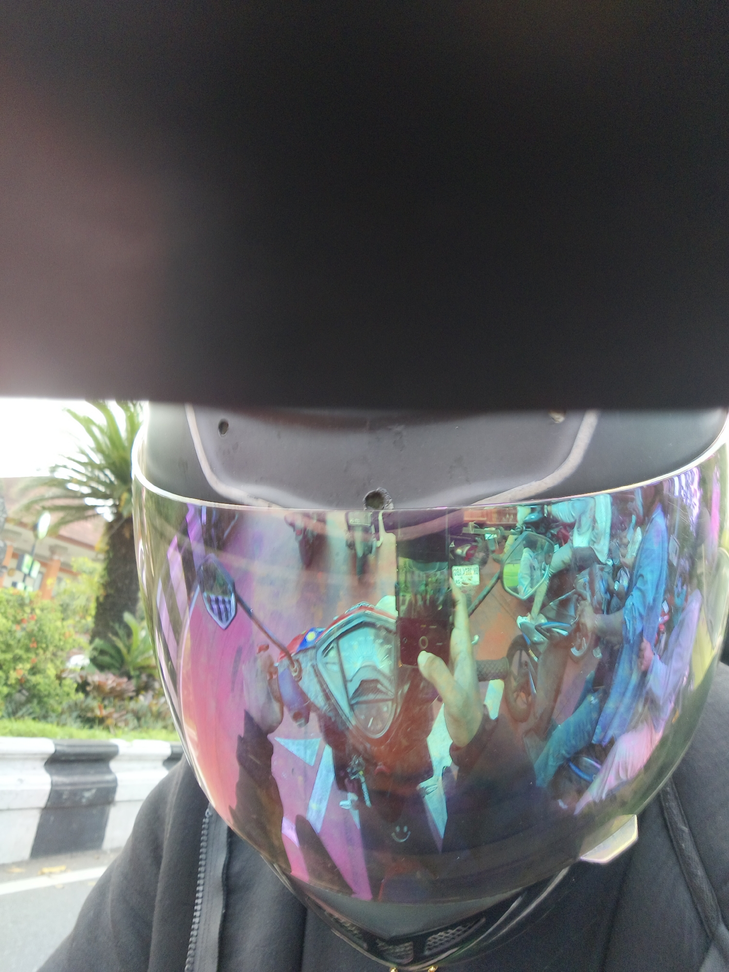 The image shows a first-person perspective of someone wearing a motorcycle helmet. The helmet's visor, which has a iridescent, rainbow-like sheen, reflects a scene of a street in Denpasar, Indonesia. In the reflection, several people can be seen on motorcycles, suggesting a busy urban environment. There are also buildings and vegetation visible in the background of the reflection. The person wearing the helmet is wearing a dark jacket. The overall setting appears to be daytime, with natural light illuminating the scene.