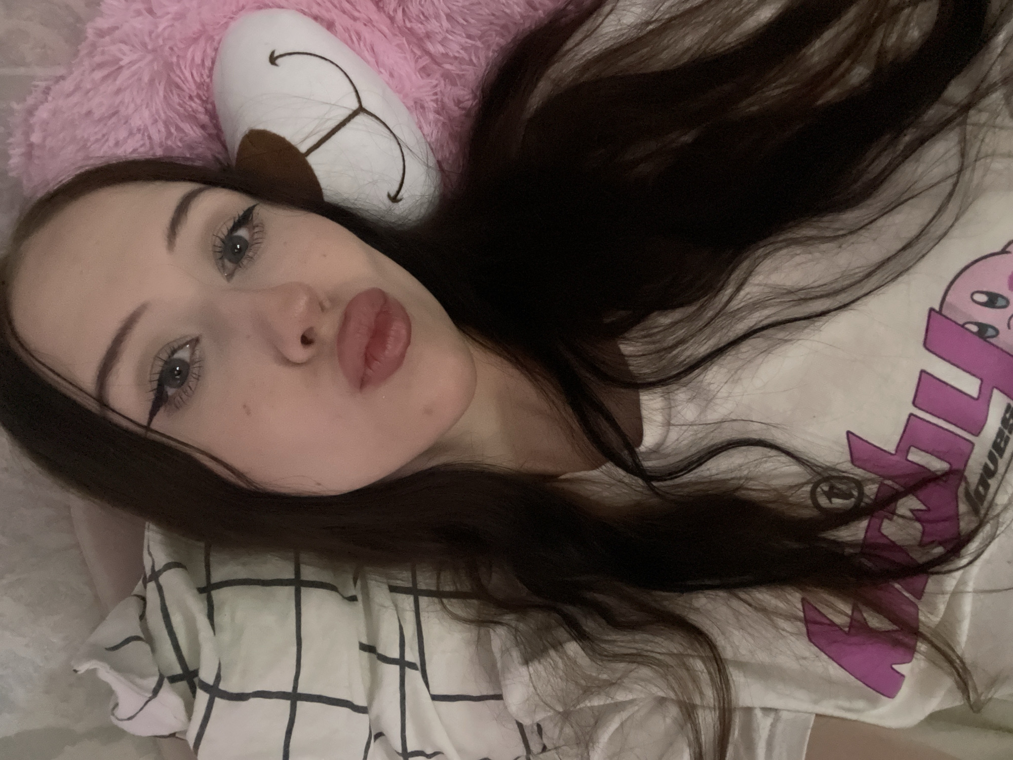 The media file is a close-up, overhead shot of a young woman lying down. She has long, dark brown hair that is spread out around her head. Her eyes are open and looking towards the camera, and she has applied eyeliner and mascara. Her lips are slightly pursed, and she appears to be wearing lipstick. She is wearing a white t-shirt with a purple graphic design on it, and a white pillow with a black and pink checkered pattern beneath her head. To her left, a pink fluffy pillow is visible. The lighting suggests it is indoors, likely in a bedroom. The overall mood is casual and relaxed. The graphic on the t-shirt appears to have some text, but it is partially obscured by her hair.
