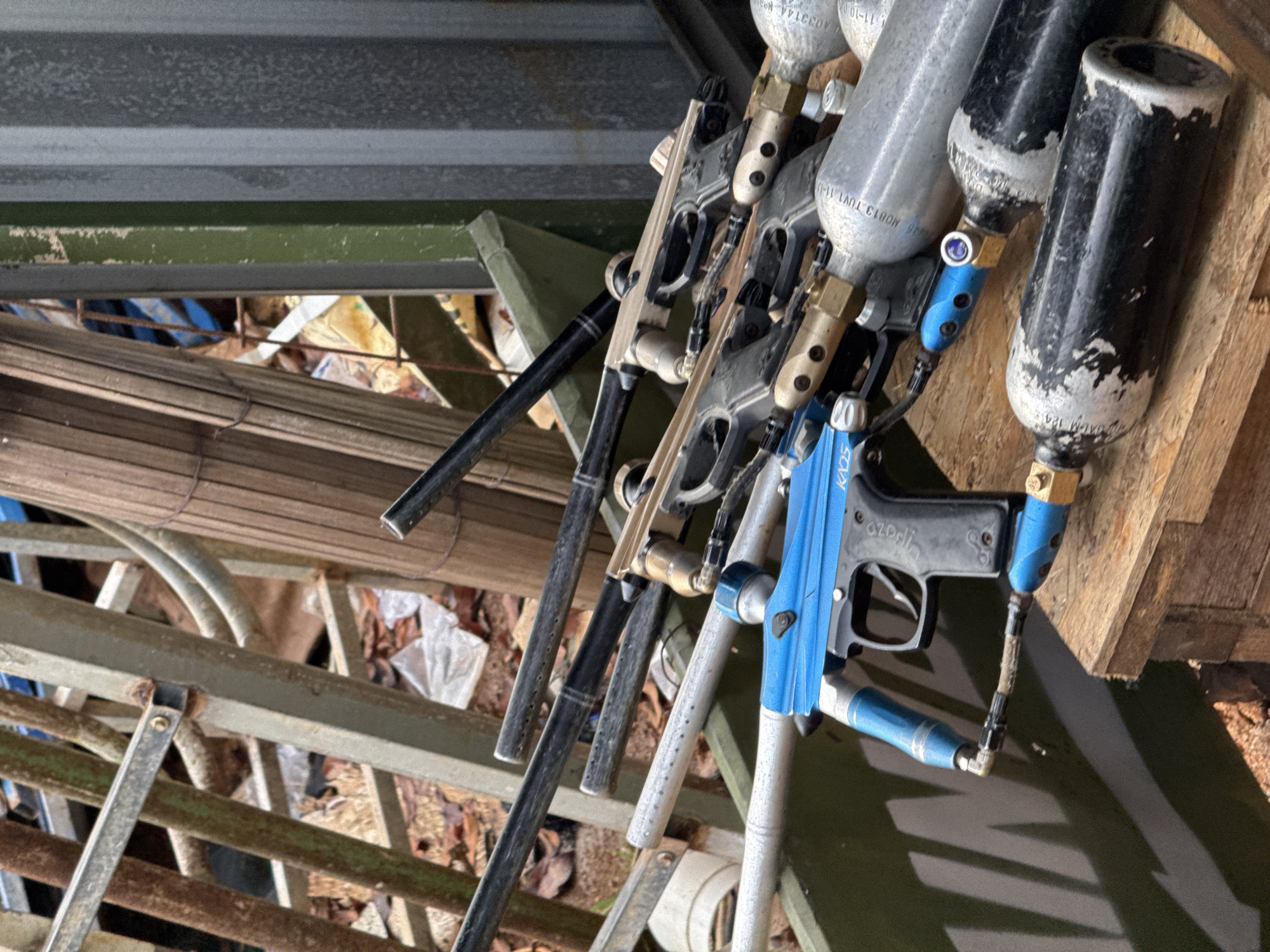 The image shows an assortment of paintball guns with attached air tanks. The guns vary in color, including black, silver, and blue. The air tanks are primarily silver and black. The background includes metal scaffolding, wood planks, and debris. The location in Kuta Selatan, Indonesia is not discernible from the image. There are no people present.