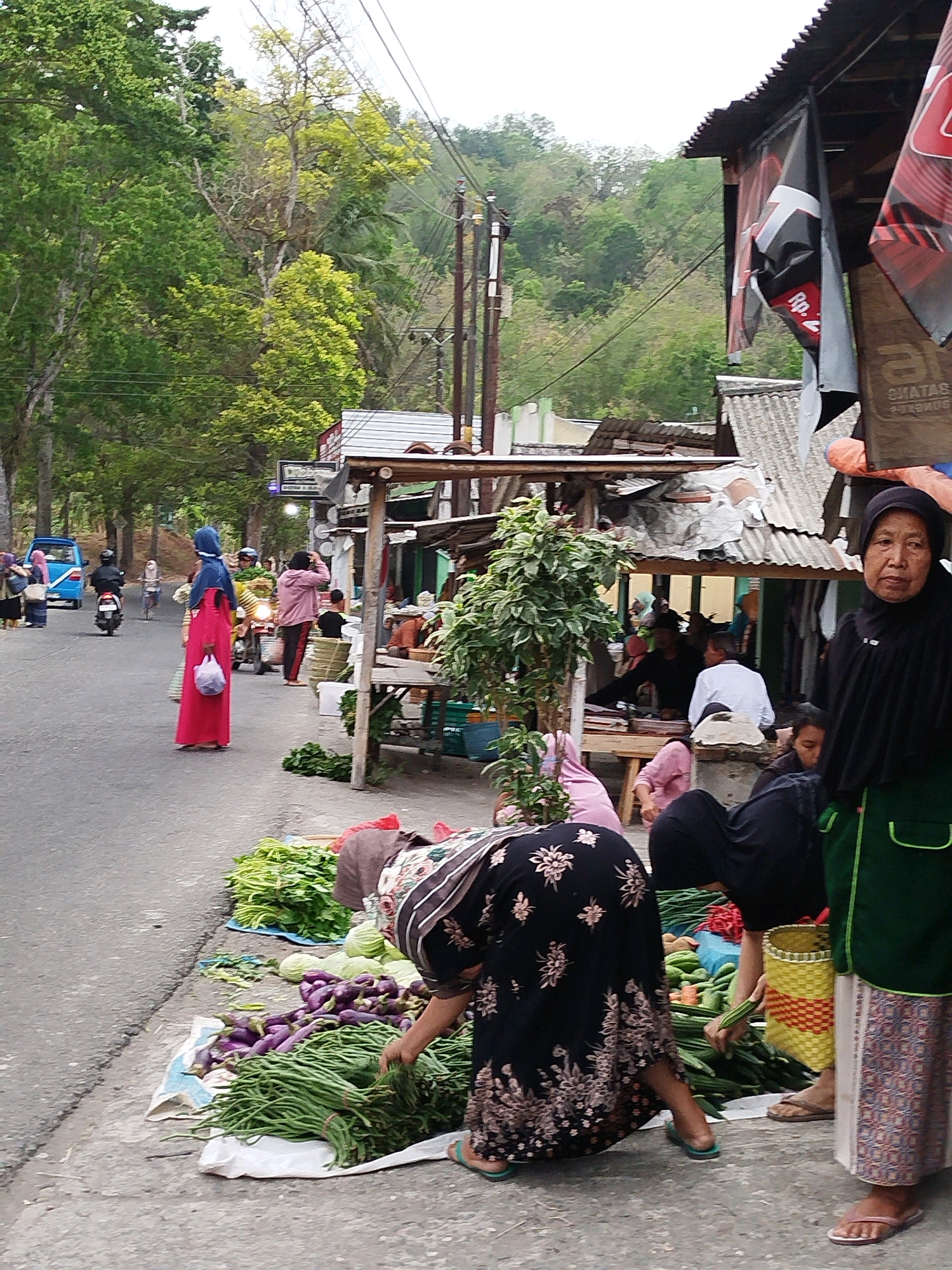 The image depicts a street market scene in Kedungwaru, Indonesia, on a paved road lined with trees and vegetation on a hillside. Several individuals are present, engaged in various activities. In the foreground, a woman wearing a black hijab and green apron stands facing the viewer, while another woman in a black patterned dress is bent over a display of long beans and eggplants on a white cloth on the ground. A yellow and red woven basket is positioned near the vegetables. Further down the street, a woman in a red dress and blue hijab is walking, carrying a white plastic bag. A blue minivan is parked on the left side of the road, with other people and motorcycles visible in the background. Stalls with corrugated metal roofs are set up along the right side of the road, some displaying banners. The overall atmosphere suggests a busy marketplace.