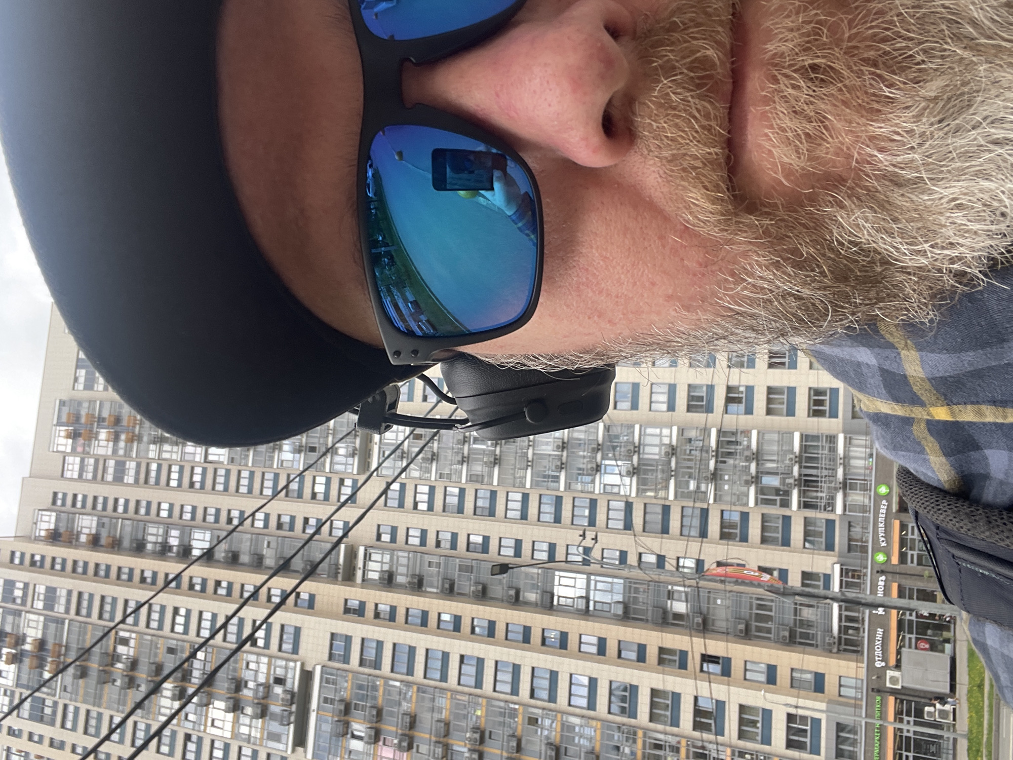 A close-up portrait of a man with a gray beard and blue sunglasses, taken outdoors in the city of Khimki, Russia. The sunglasses reflect an urban environment, specifically a tall apartment building with numerous windows and air conditioning units, as well as street signs visible in Russian. The man is wearing a black head covering and headphones. His face is positioned in the foreground against the backdrop of the apartment building and the street. The perspective is tilted, with the building appearing below the man's face. The man is also wearing a plaid shirt.