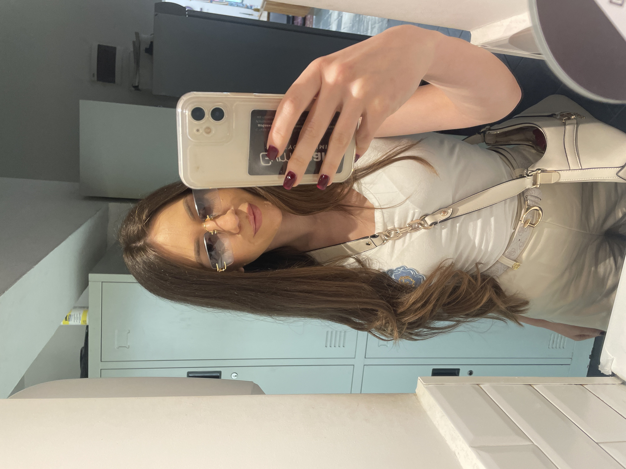 A woman is taking a selfie in a bathroom with lockers visible in the background. She is wearing sunglasses, a white top, and a crossbody bag. Her nails are painted a dark color. The location appears to be an indoor space, possibly a school or gym, with tiled walls and a vanity countertop. There is no concrete evidence to place this image specifically in Sofia, Bulgaria.
