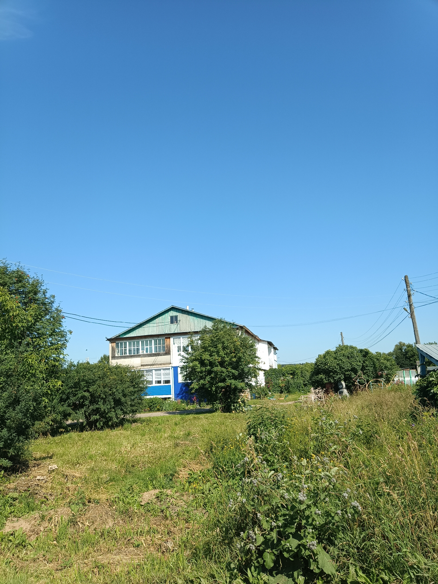 The image depicts a two-story building with a green, sloped roof, possibly a house, situated in a rural or semi-rural area. The building has white and blue sections, with a balcony on the upper floor featuring white framed windows. It is surrounded by lush green trees and dense vegetation, including tall grasses and flowering plants. A wooden utility pole with electrical wires is visible to the right of the building. The sky is a clear, bright blue. The scene appears to be a daytime shot, under sunny conditions, likely in a village or dacha settlement. The specific location in the city of Посад, Russia, cannot be determined from the visual information alone.