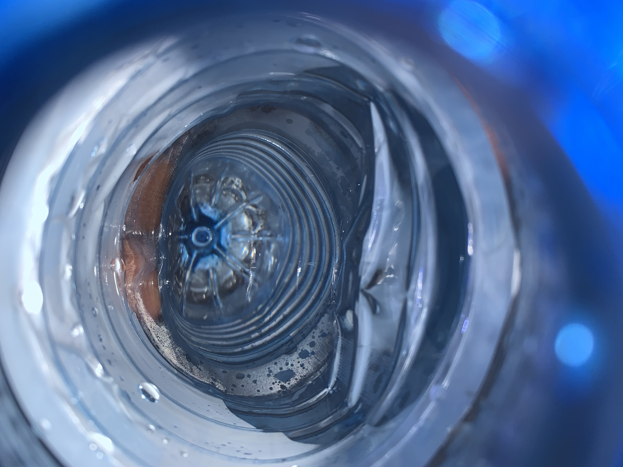 The image depicts the interior of a blue-tinted plastic water bottle. The perspective is from inside the bottle, looking outwards. The bottle is partially filled with water, as evidenced by the visible water droplets and the waterline. The bottom of the bottle is visible in the center, with a star-shaped indentation. The image is taken at close range. The bottle shows signs of wear and tear. It is impossible to determine the location as Jos, Nigeria from this image.