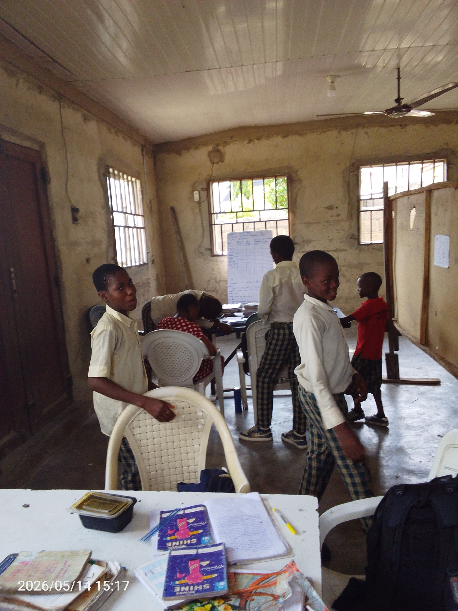 Inside a classroom in Lokoja, Nigeria, several children are present. One boy in a khaki shirt and plaid pants stands in the foreground, facing the camera. Another boy, also in a khaki shirt and plaid pants, is walking near the center of the room. A third boy, wearing a red shirt and dark shorts, stands near the back. Other children are seated at desks. The classroom has several barred windows, a wooden ceiling, and a ceiling fan. A table in the foreground holds books, papers, and a black container. The timestamp "2026/05/14 15:17" is visible.
