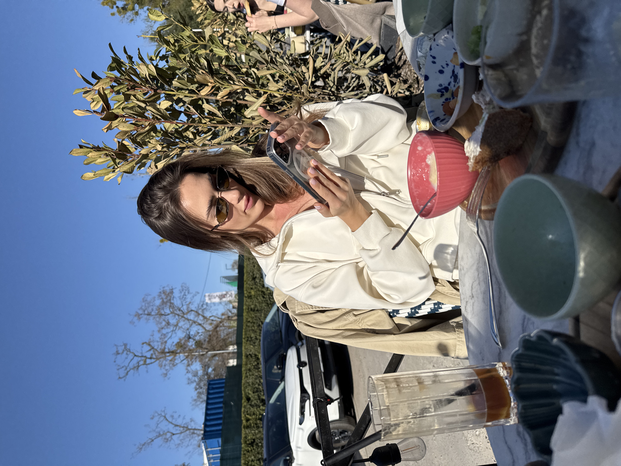 The image shows a woman with sunglasses, holding a smartphone in a clear case, at an outdoor table filled with dishes and cups. She is wearing a white hoodie and a tan jacket. In the background, there is a car and trees silhouetted against a blue sky. The location is in Warsaw, Poland, indicated by the signs visible behind her.