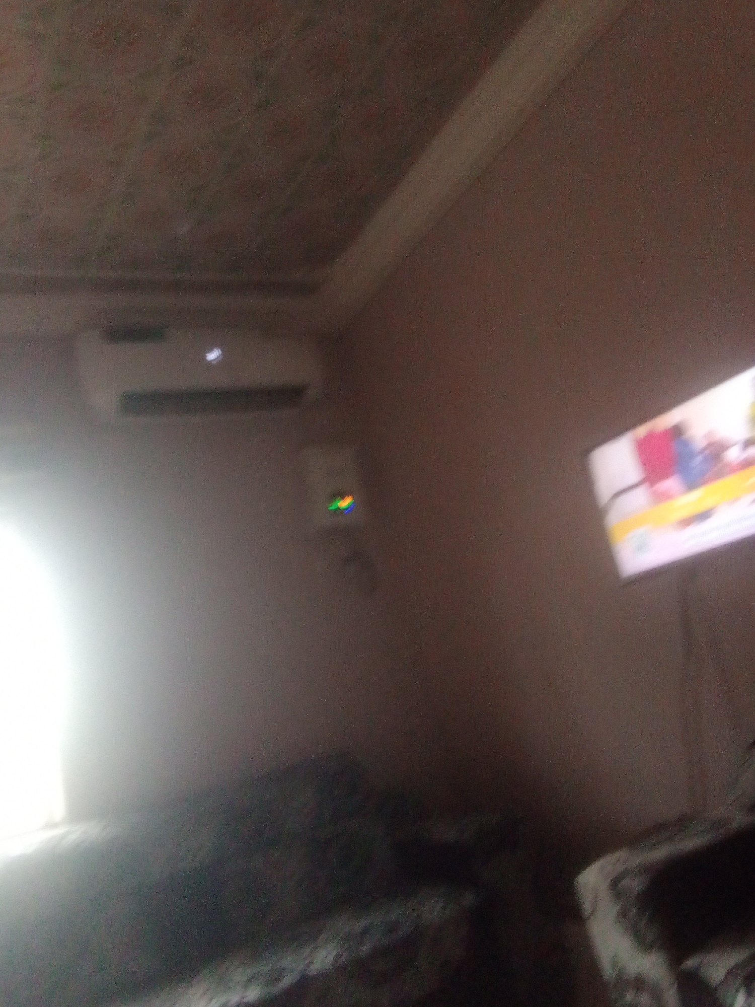 The image shows an indoor scene, likely a living room, in Jalingo, Nigeria. A white air conditioning unit is mounted high on a light brown wall. Below the AC unit, a small device with a rainbow-colored light is visible. To the right of the AC unit, a television screen displays a colorful image, possibly from a program or advertisement, with shapes that suggest people or objects.
The lighting in the room appears dim, with a bright, hazy light source to the left, possibly a window. The ceiling has a textured pattern. In the foreground, a dark, upholstered surface, possibly a sofa or bed, is visible, partially obscured by shadows and a bright light flare. The overall impression is of a room at dusk or night, with artificial light sources dominating the illumination. No text is clearly legible in the image.