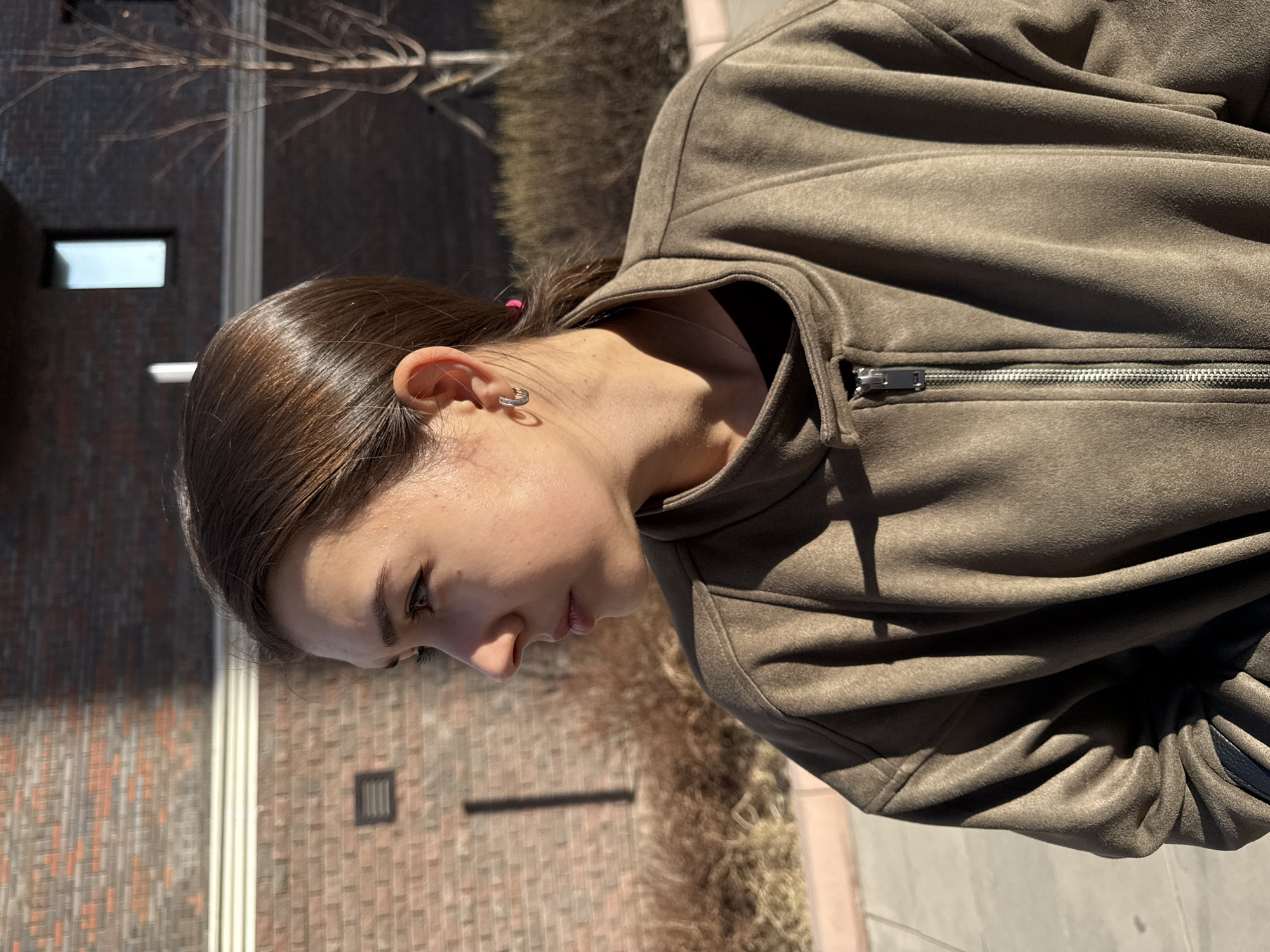 A woman with brown hair pulled back is shown in a profile shot, wearing a tan jacket. She is standing outside, with a brick building and dry brush visible in the background. A small silver hoop earring is in her left ear. The location is not discernible as Krasnoyarsk, Russia.