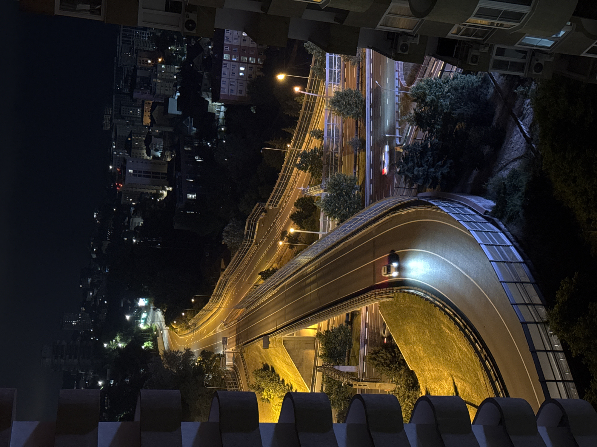 This is a nighttime aerial view of a curved highway in Sochi, Russia. The highway is illuminated by streetlights, casting a warm glow on the asphalt. On the highway, one car is visible, with its headlights on and illuminating the road ahead. The car is on the right lane of the highway, moving away from the viewer. The highway is bordered by a metal fence and a grassy embankment. In the background, a city skyline is visible, with numerous buildings illuminated by lights. Some of these buildings appear to be residential, with windows lit up. The overall scene depicts a modern urban environment at night, with the highway serving as a prominent feature. The presence of balconies on the right side of the image suggests that the photograph was taken from a high vantage point in a multi-story building.