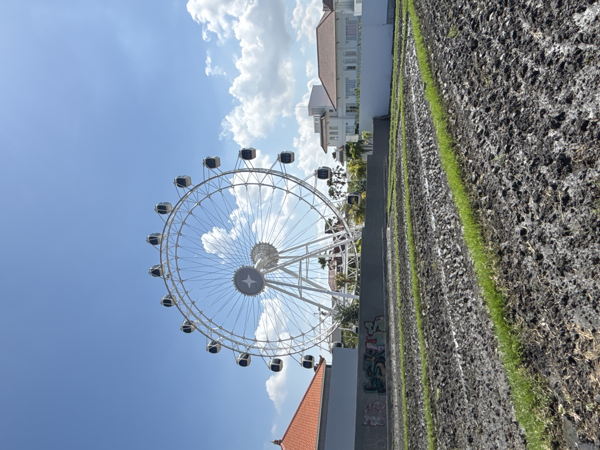 An outdoor, full-shot photograph captures a Ferris wheel standing tall against a bright blue sky with scattered white clouds. The Ferris wheel is predominantly white and features numerous gondolas. In the foreground, a muddy field with rows of young green shoots stretches across the frame. Behind the field, a dark gray wall is visible, adorned with graffiti. To the left of the Ferris wheel, a building with a red-tiled roof and a gray facade is present. To the right, several white buildings with red-tiled roofs, characteristic of North Kuta, Indonesia, are situated. The scene appears to be a leisure area within a city, possibly an amusement park or entertainment complex, with elements of urban development contrasted against agricultural land.
