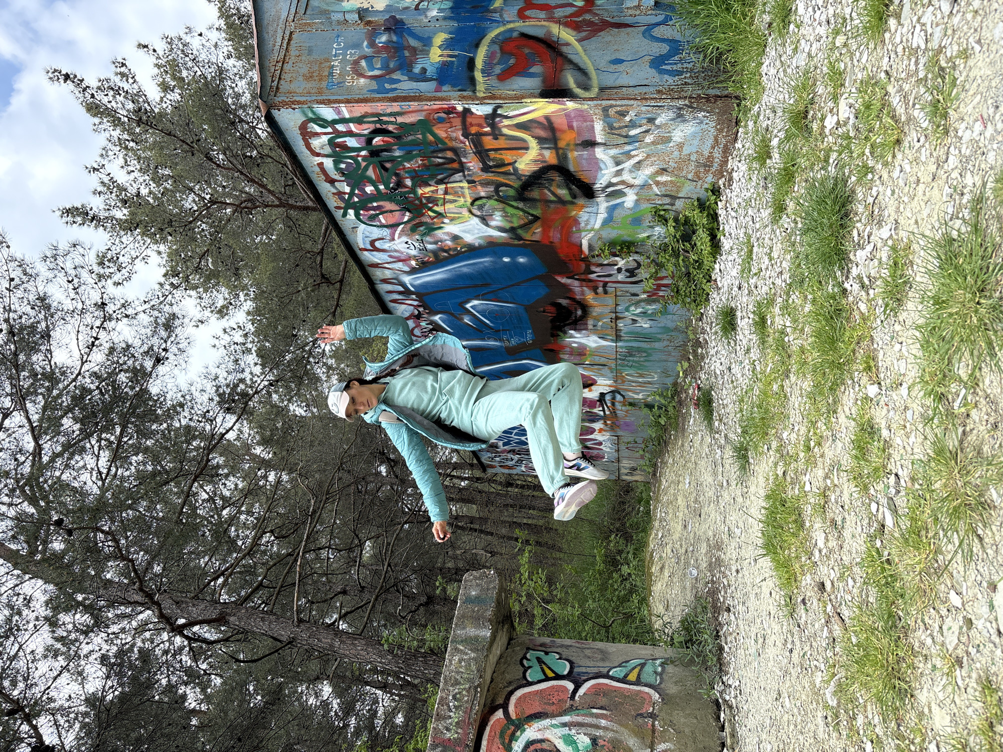 This image, rotated 90 degrees counter-clockwise, captures a dynamic outdoor scene in Gelendzhik, Russia, featuring a person in mid-air against a backdrop of graffiti and nature.
The main subject is a person, likely a woman, dressed in a light mint green tracksuit, a light blue quilted jacket, and a white baseball cap. She is shown in a suspended pose, with arms extended horizontally and legs slightly bent, as if jumping or floating. Her expression appears calm.
The setting combines elements of urban decay and natural landscape. Behind the person is a tall, extensively graffiti-covered metal wall or structure, adorned with a chaotic mix of vibrant spray paint in colors such as blue, red, yellow, black, and white, over a rusted light blue base. Visible near the top of this wall are Cyrillic characters reading "ПРОДАЕТСЯ 95-0-63," which translates to "FOR SALE 95-0-63." To the left and above, a dense canopy of tall pine trees with dark needles fills much of the frame, hinting at a forest or wooded area.
The ground below is composed of small, light-colored pebbles mixed with patches of green grass. In the foreground, a concrete block, also marked with graffiti, is partially visible. The sky, seen through the trees, is overcast or partly cloudy, suggesting a daytime shot. The overall impression is one of playful energy against a textured, industrial-natural environment.