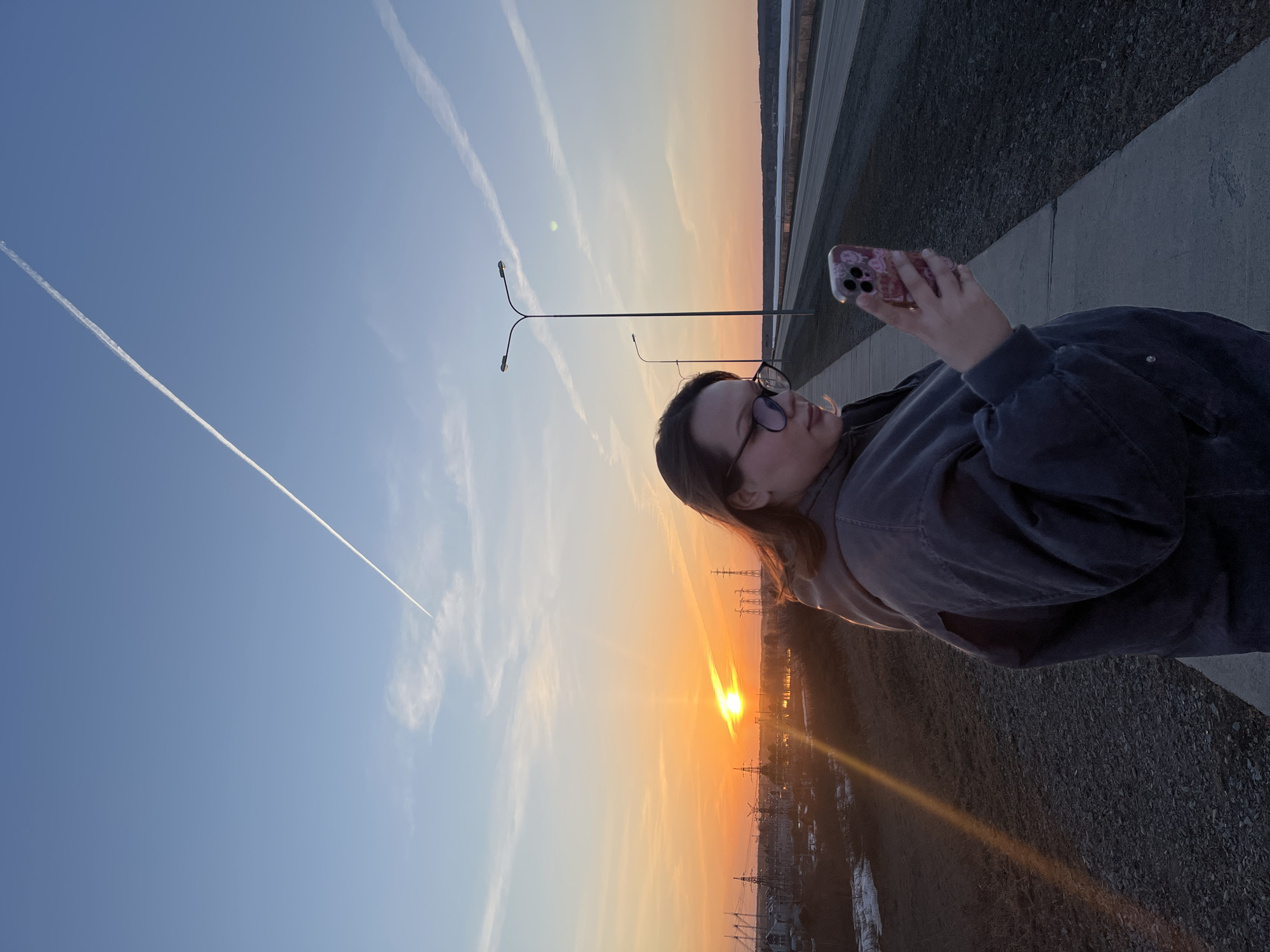A medium shot shows a woman standing on a gray concrete walkway taking a photo of a vibrant orange sunset in Chaikovsky, Russia. She is positioned to the right, facing towards the right, with her head angled slightly downward as she looks at her phone. She wears glasses and a dark gray jacket.
The background features a cityscape with numerous electrical towers and power lines silhouetted against the setting sun. The sky transitions from blue to orange, displaying contrails. The left side of the image shows the blue sky and the right side shows the darkening ground covered in gravel. Two street lights stand nearby.