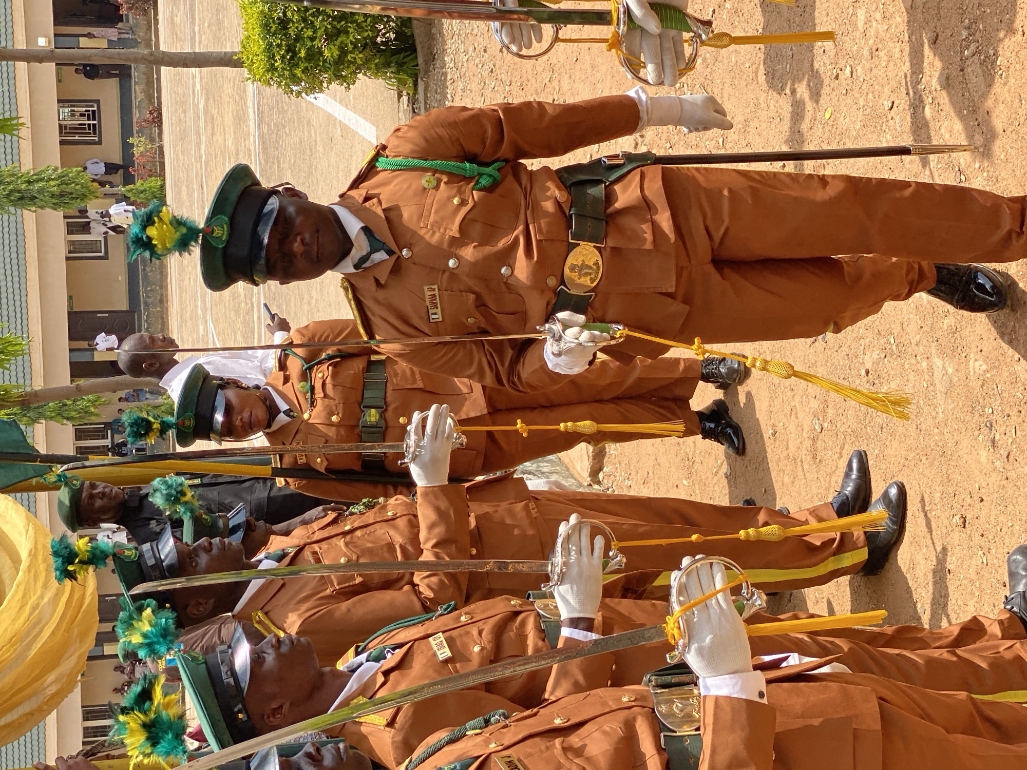 The image depicts a group of individuals, likely military or police personnel, in uniform. They are standing in formation outdoors, on what appears to be a parade ground or a similar open area with sandy soil and some sparse vegetation. The individuals are dressed in matching tan uniforms with green accents, including green sashes and hats adorned with green and yellow plumes. They are all holding ceremonial swords, some with white gloves.
The formation suggests a formal event, possibly a parade, ceremony, or inspection. The subjects are facing forward, seemingly attentive. The time of day appears to be daytime, with shadows visible on the ground, indicating sunlight. The weather seems fair, suitable for an outdoor event.
In the background, a building with a light-colored facade and a tiled roof is visible, along with some trees and other people in civilian attire. There is a glimpse of a yellow banner or flag on the left side of the image.
One individual's uniform has a name tag that reads "I. SARAWANA AP". The presence of uniforms, swords, and a formal formation points to an official occasion. The overall atmosphere conveyed is one of discipline and solemnity. The location context of Kaduna, Nigeria, suggests this could be a Nigerian military or police event.