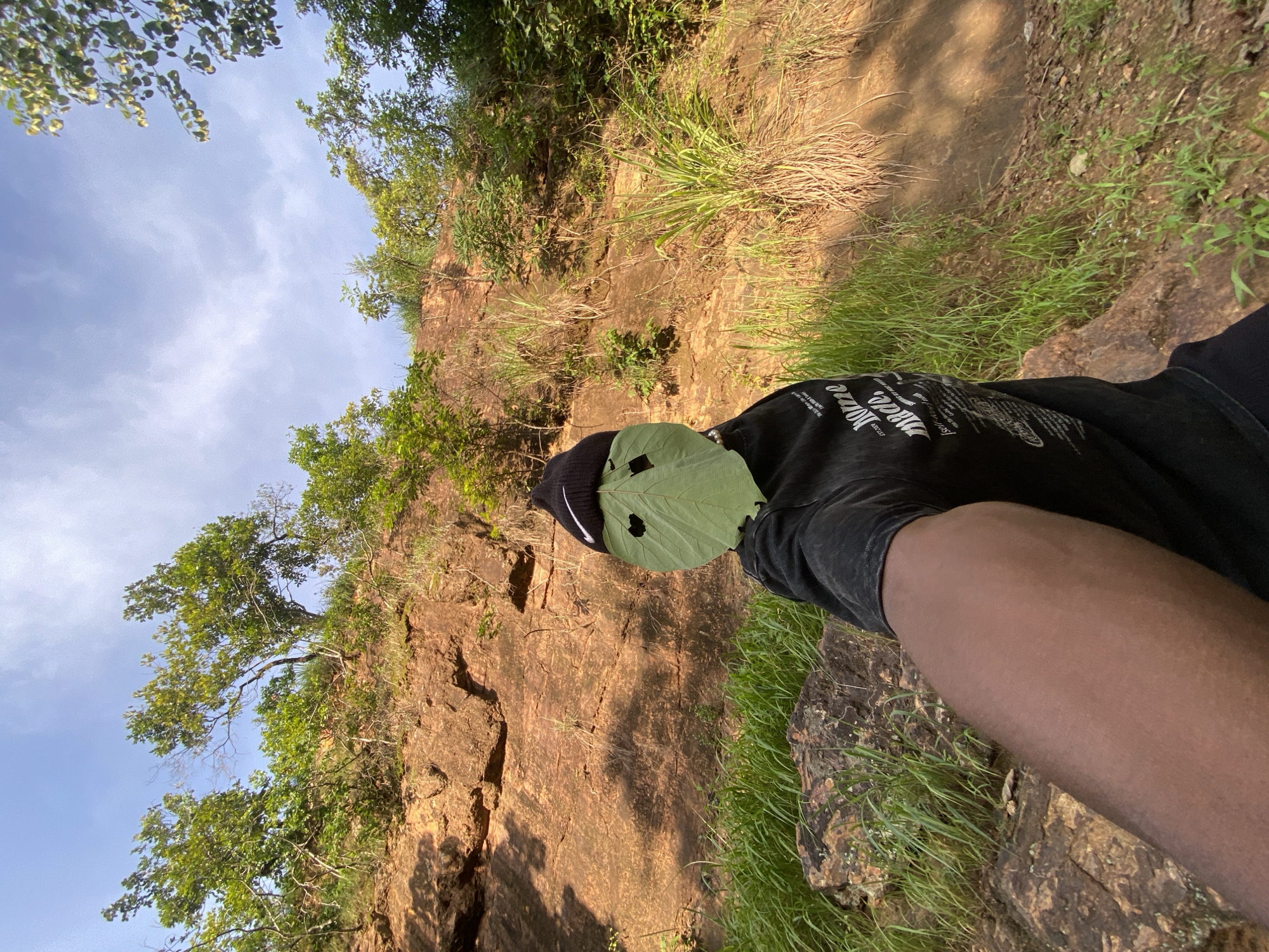 A person is standing on a rocky, grassy slope outdoors, holding a camera to take a selfie. The person is wearing a black beanie with a white Nike logo and a black t-shirt. A large green leaf is placed over the person's face, with cutouts for eyes and a mouth, creating a mask. The background shows a steep, reddish-brown rocky cliff face covered in green foliage and trees. The sky is blue with scattered white clouds. The scene appears to be in a natural, undeveloped area, possibly a park or hiking trail. The lighting suggests it is daytime, likely late afternoon due to the warm tones of the rock. The environment does not provide specific clues to pinpoint a precise location within Abakaliki, Nigeria, but it depicts a natural landscape consistent with areas found in the region.