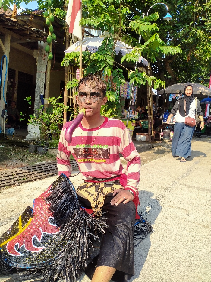 The image depicts a daytime scene on a paved surface in Sepat, Indonesia. In the foreground, a male individual is seated, looking directly forward. He has light-colored hair and his face is marked with dark paint around his eyes, nose, and cheeks. He wears a red and white horizontally striped long-sleeve shirt with a graphic print on the chest, dark trousers, and a patterned fabric wrapped around his waist. A curved, purple eggplant is held in his mouth. Beside him on the ground is a large, elaborate object resembling a traditional Kuda Lumping (Jaranan) horse puppet, featuring black, red, and yellow patterns with black fringe.
In the mid-ground, an adult female wearing a black hijab, white shirt, blue wide-leg pants, and carrying a brown shoulder bag walks from right to left. The background features various elements: on the far left, a light-colored building with a visible porch and a blue hose; on the right, several small vendor stalls or shops with merchandise displayed under umbrellas and dense tree foliage. A papaya tree bearing green fruit is visible on the left, and an Indonesian flag is displayed on a pole. The scene is illuminated by bright sunlight, casting distinct shadows.
