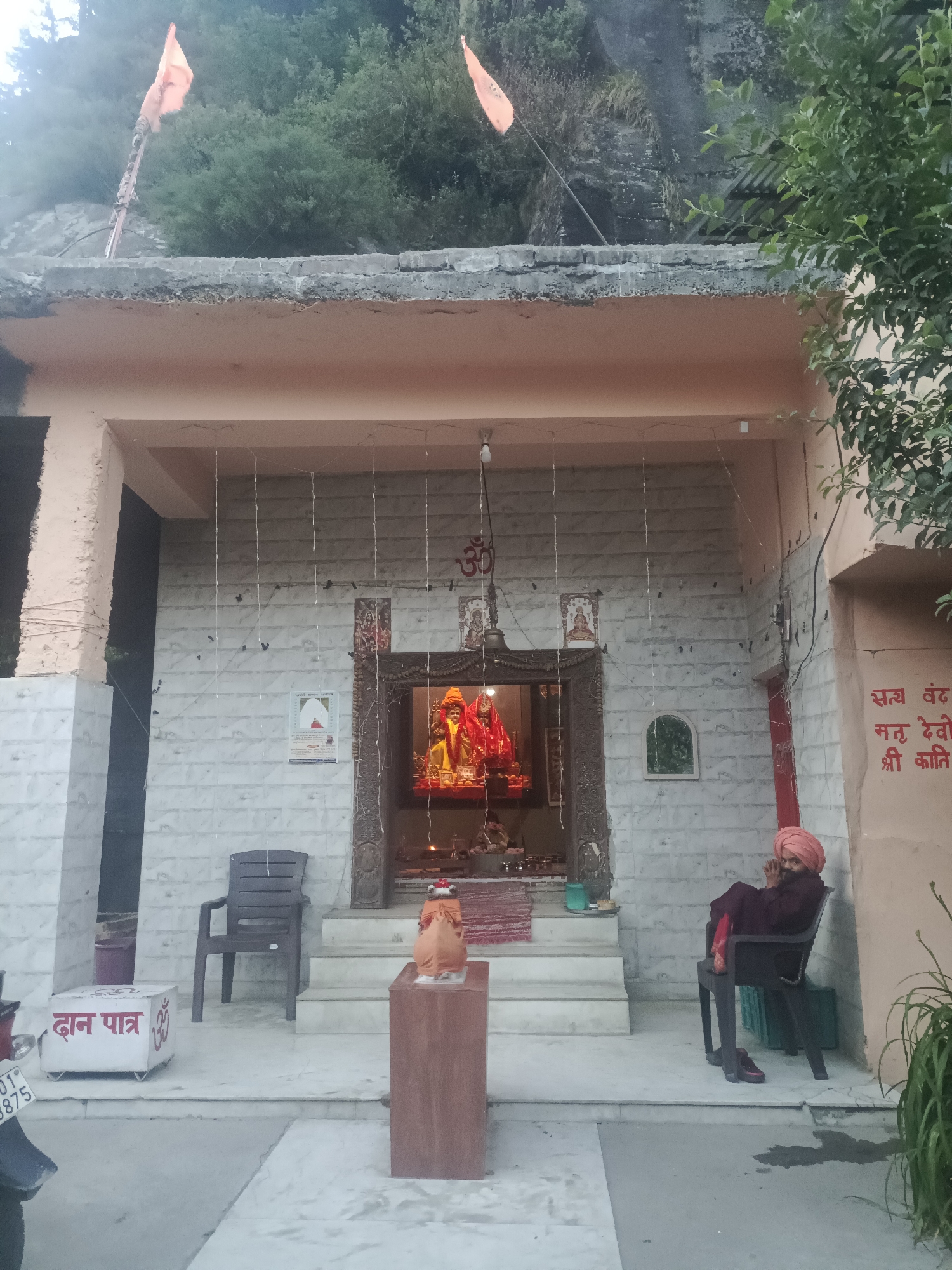 This is a medium shot, eye-level photograph of a small temple entrance. Two orange flags are visible on poles extending from behind the temple roof, against a backdrop of trees and a rocky incline. The temple facade is painted a pale peach color with a concrete roof. The main entrance is recessed and has a white tiled interior wall. Decorative white lights are strung across the ceiling of the entrance. Inside the shrine, two figures in red and yellow robes are visible, appearing to be deities. To the right of the entrance, a man wearing a burgundy robe and an orange turban is seated in a black plastic chair. He is facing left, with his hands clasped in front of his face. To the left of the entrance, a white donation box with red Hindi text and an Om symbol is visible, alongside a dark gray plastic chair. A red rug is laid out on the steps leading into the shrine. A small, framed picture of a seated person is affixed to the wall to the left of the shrine entrance. A larger framed poster with Hindi text is on the wall to the right of the shrine entrance. The scene appears to be outdoors, in a hilly or mountainous region.
