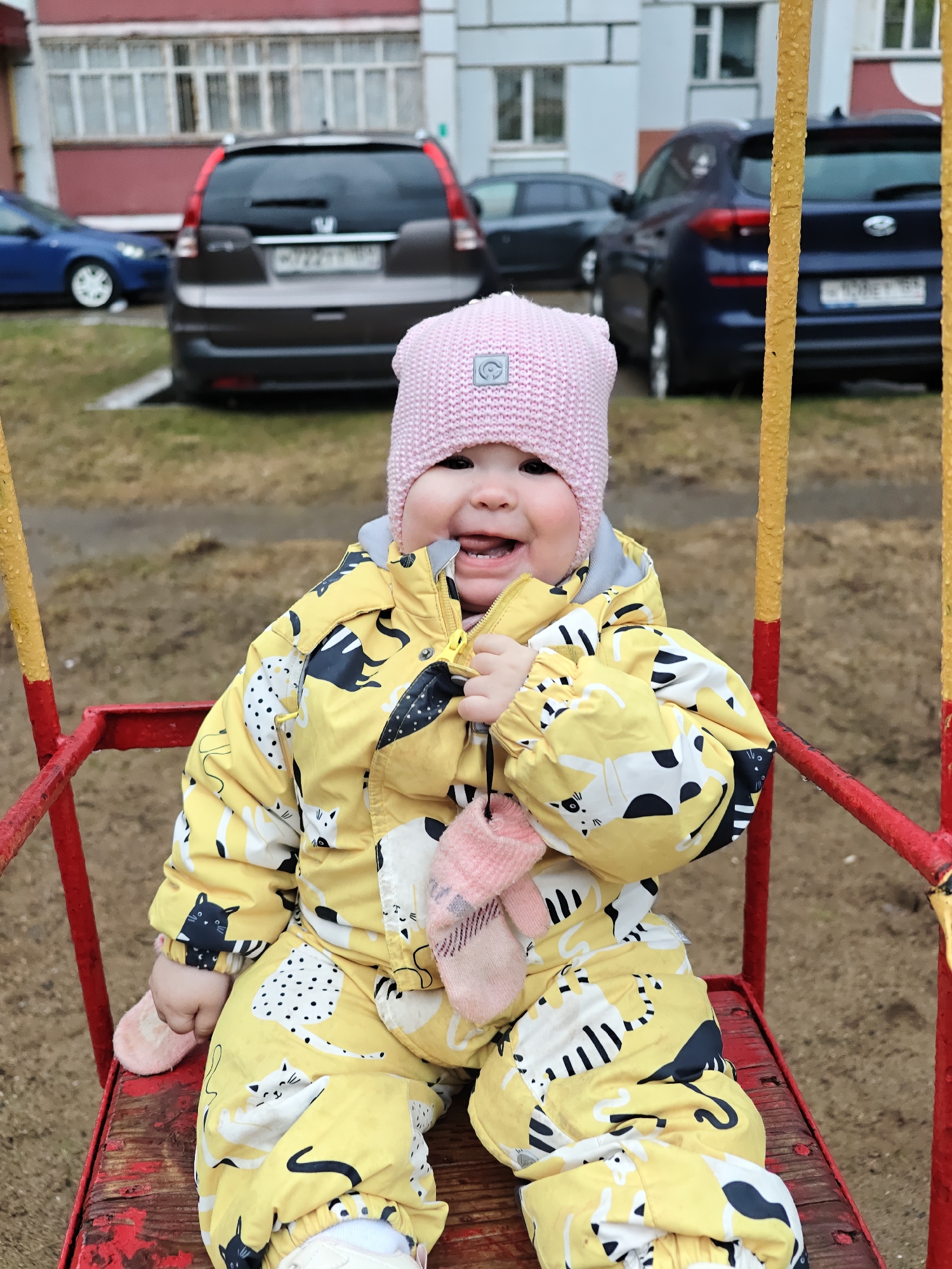 A young child, possibly a toddler, is seated on a red swing at a playground. The child wears a yellow snowsuit with a cat print, and a pink knit hat. The child is smiling and holding a pink glove near their mouth.
In the background are several parked cars and a multi-story building with windows. Based on metadata, this photograph was taken in the city of Chaikovsky, Perm Krai, Russia.