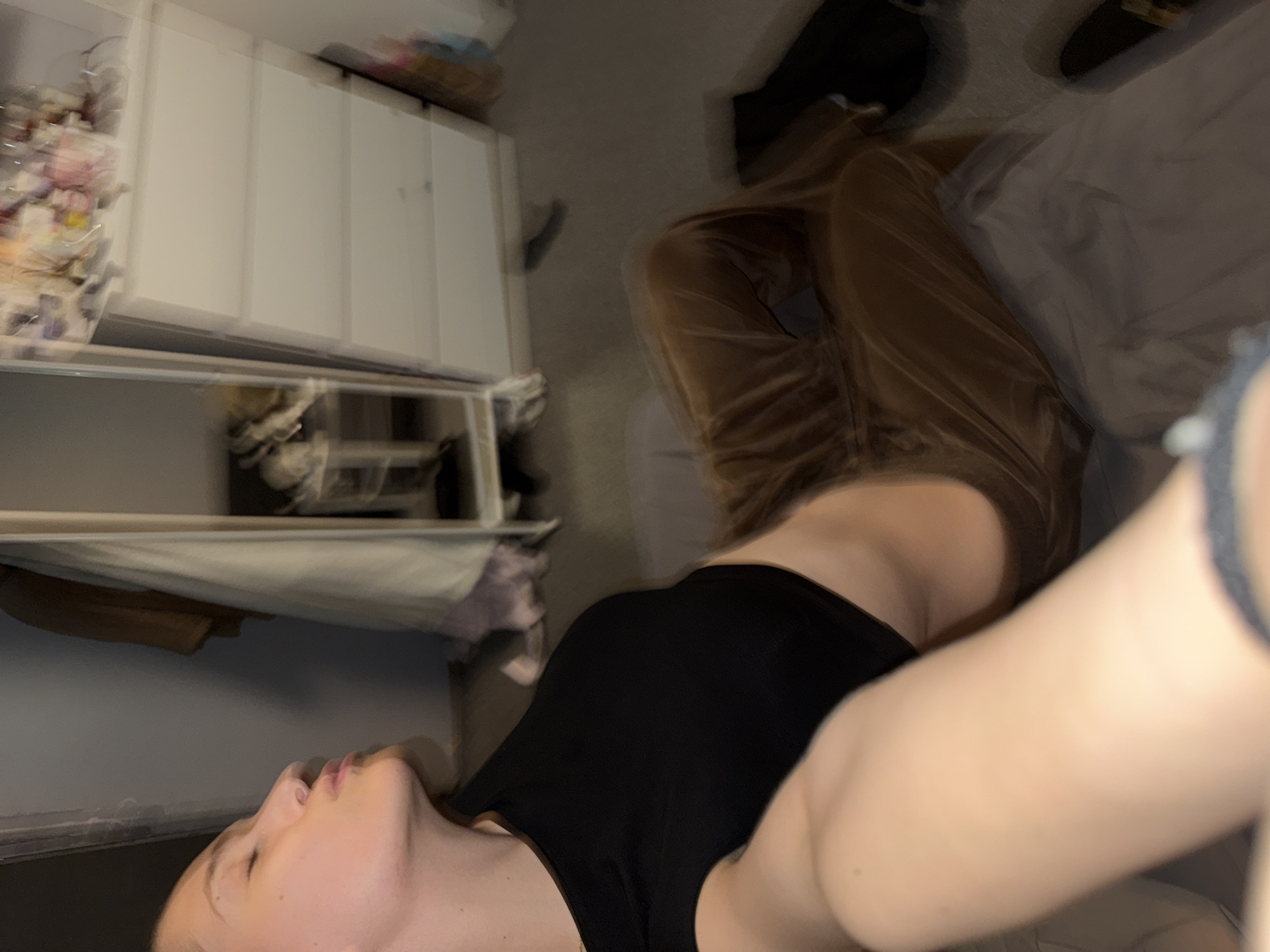 A person is lying down, facing upwards, wearing a black tank top and brown pants. Part of their face and arm are visible. The environment consists of a room with a white cabinet, shelves, and personal belongings. The location is unspecified; there is no information indicating it is in Sofia, Bulgaria.