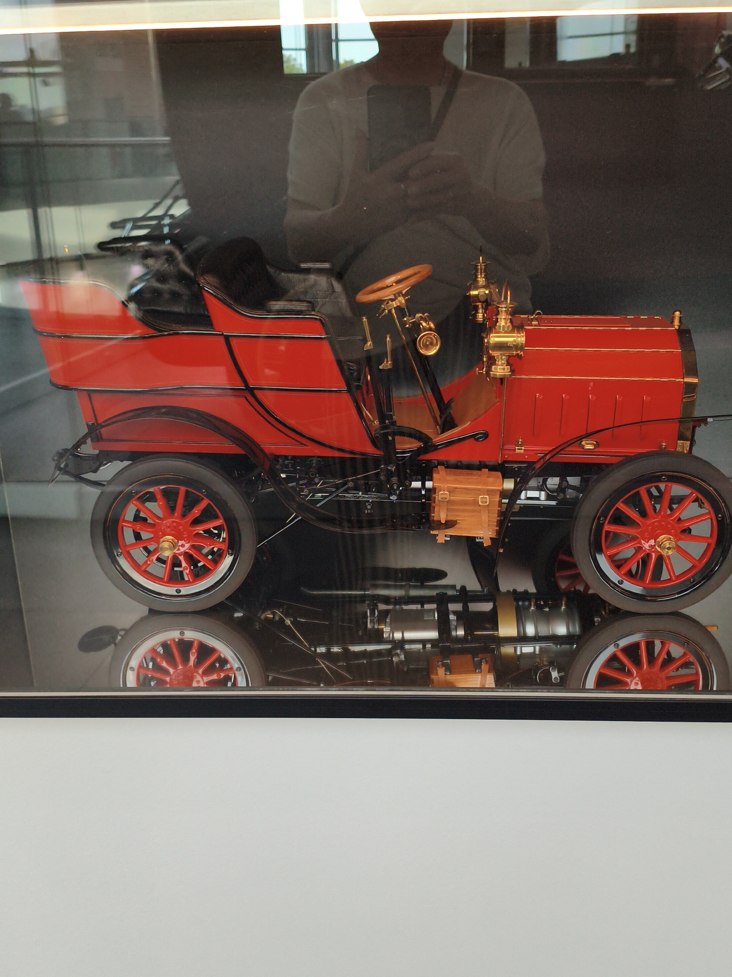 The image displays a vintage red automobile model with intricate brass detailing, positioned in a display case. The model features spoked red wheels and a wooden box on its side. A person, seen in reflection and holding a phone, is visible behind the car, appearing to be in a museum or exhibition space. The setting suggests an indoor environment, potentially the Audi Forum Ingolstadt, given the context of vintage automobiles. The car's reflection is clearly visible on a polished surface below it.
