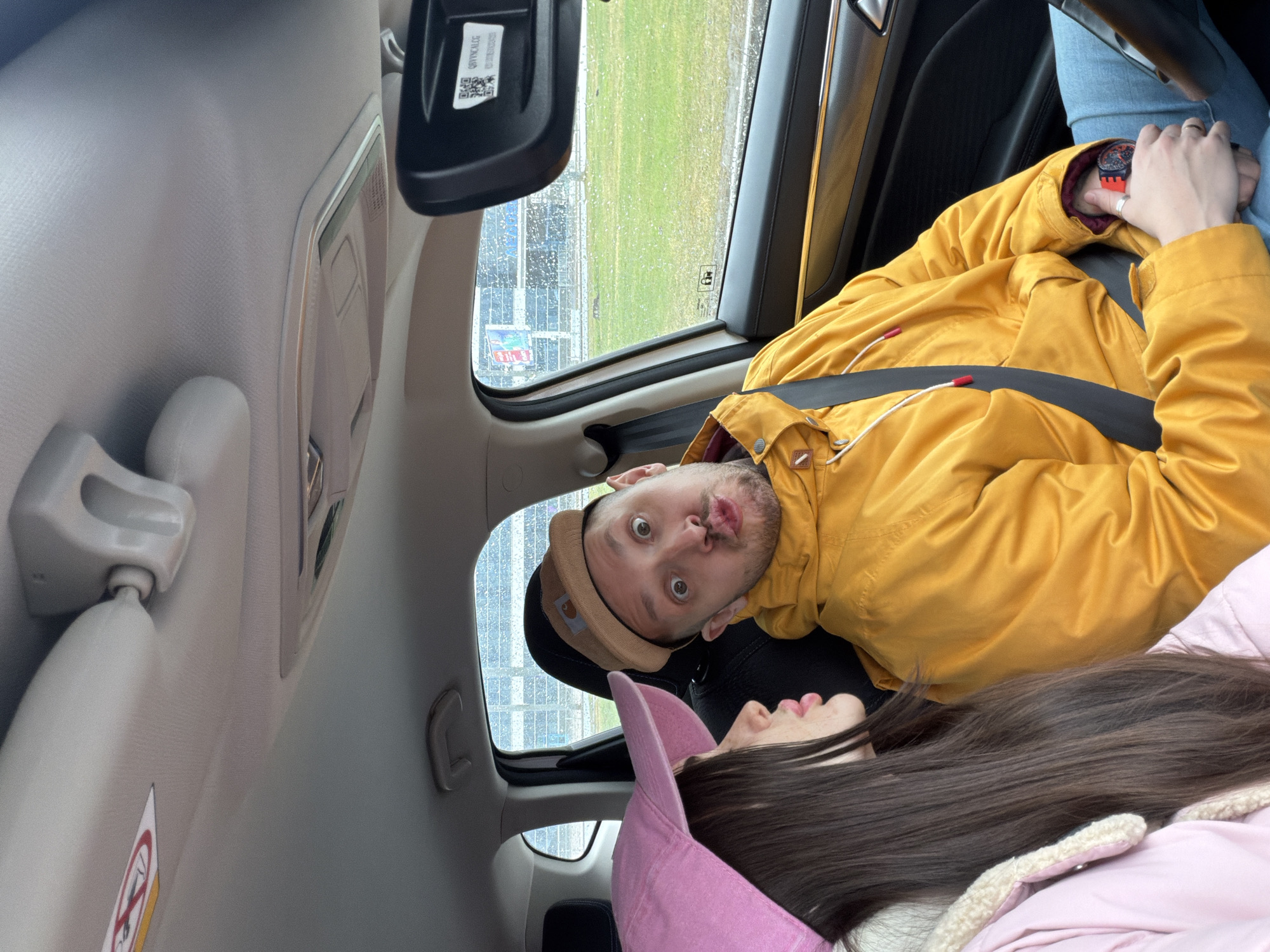 Inside a car, a man and a woman are visible. The man, wearing a tan hat and a yellow jacket with a seatbelt, looks directly at the camera with a surprised expression. The woman, wearing a pink hat, is lying down. Rain streaks the window, and outside is a view of a grassy area with what appears to be a building in the distance. The location appears to be near the Saint Petersburg Ring Road in Russia.