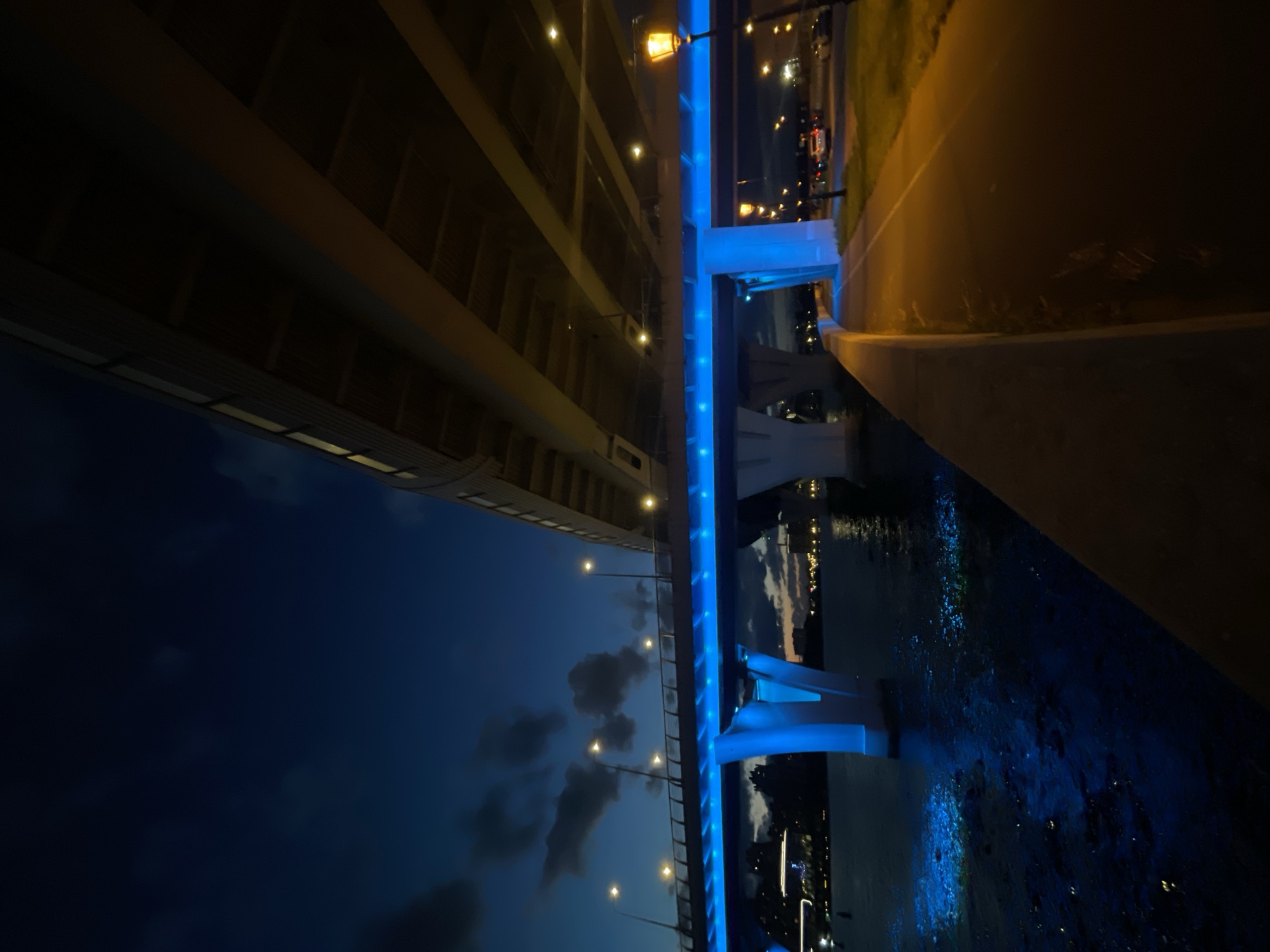 This is an outdoor, low-angle, medium shot of a bridge illuminated with blue lights at night. The bridge spans over a body of water, which reflects the blue lights and appears to have some bioluminescence or other light sources within it. On the right side of the image, there is a pathway or sidewalk next to the water, illuminated by orange streetlights. Beyond the pathway, there is a road with a few cars visible. The sky above is dark blue with some scattered clouds, suggesting it is either dusk or night. The scene is serene and modern, with the contrast between the blue bridge lights and the warm streetlights creating a visually interesting atmosphere. The location is the city of Unknown, Unknown.