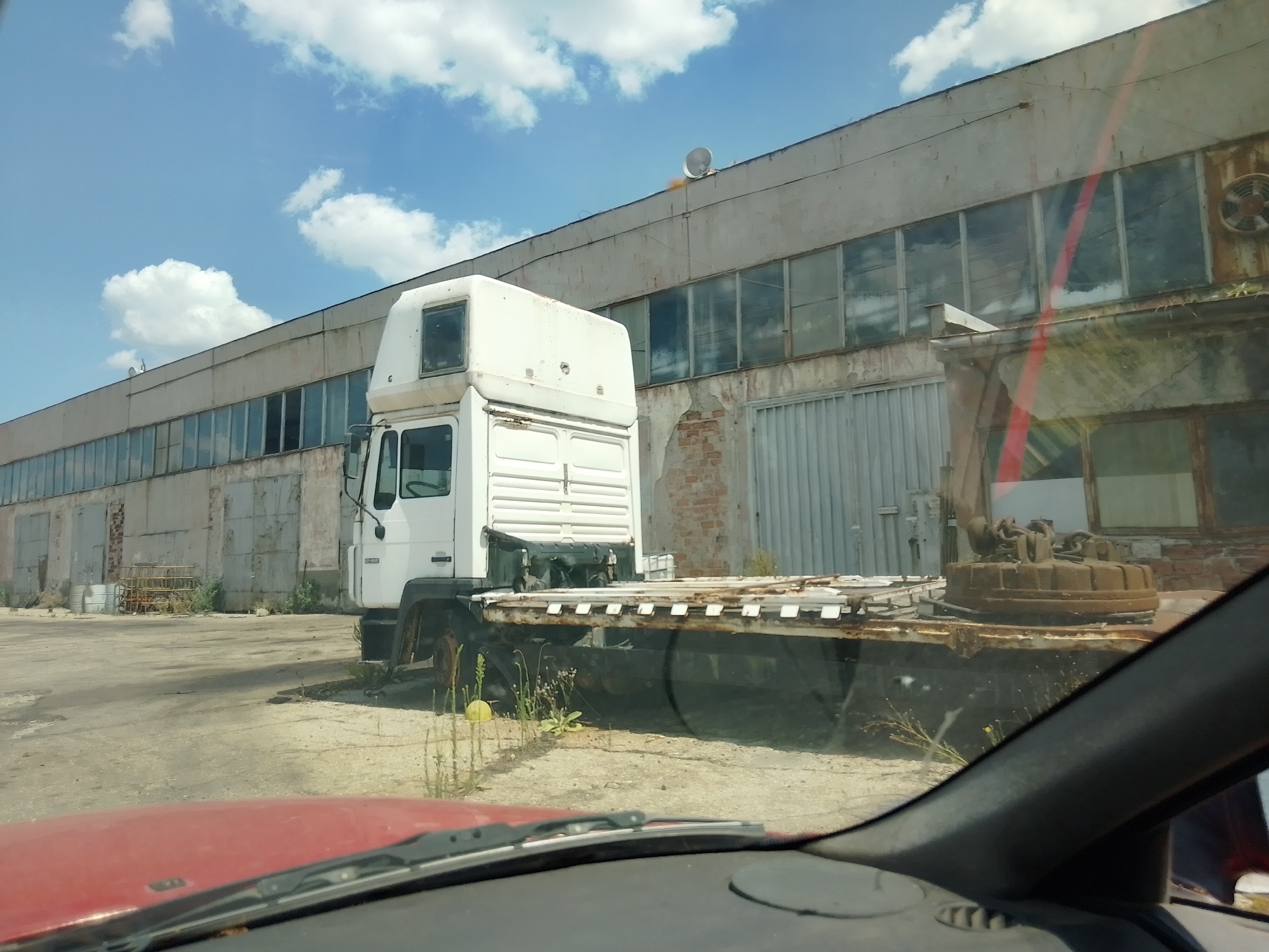 The image depicts a flatbed truck with a white cab parked in front of a long, industrial-style building. The truck's flatbed is empty except for a large, round, rusty object that appears to be a magnetic crane attachment, secured with chains. The building has a row of tall, rectangular windows with some broken panes and is constructed from concrete or a similar material. A corrugated metal door or wall is visible on the right side of the building. The ground in front of the building is a dirt and gravel lot, with some sparse vegetation growing in patches. The sky is blue with scattered white clouds. The scene appears to be an outdoor industrial or storage area. The foreground shows a portion of a vehicle's interior, including a red dashboard and a windshield wiper.