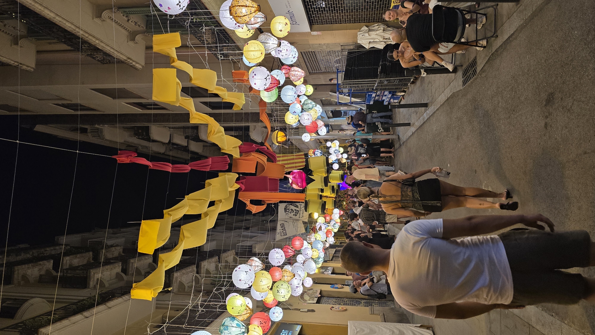 An exterior, vertical, medium shot captures a street scene in Barcelona, Spain at night, adorned with elaborate decorations for what appears to be a festival. The street, lined with multi-story buildings, is illuminated by a variety of colorful paper lanterns, predominantly white, yellow, red, and blue, suspended overhead. Festive fabric streamers, in yellow, red, and orange, are also draped across the street, creating a vibrant canopy. Numerous people are present, walking along the street, socializing, and seated at tables outside businesses. In the foreground, a man with his back to the camera, wearing a white t-shirt and dark shorts, walks away from the viewer. To his right, a woman in a black dress and sandals walks ahead of him, also facing away. On the right side of the street, several people are seated at tables, some appearing to be dining or conversing. The street is paved with asphalt, and visible on the sides are shop fronts, awnings, and streetlights. The overall atmosphere is lively and festive, indicative of a public celebration or event.