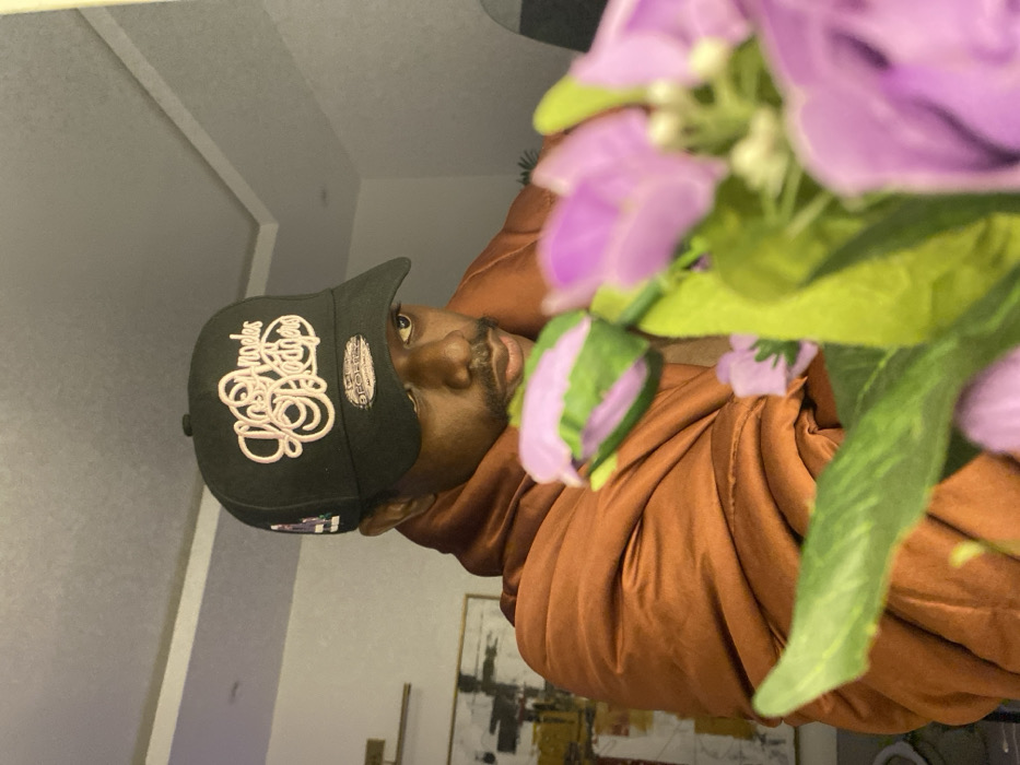 A man wearing a black baseball cap with white embroidery and an orange hooded jacket is visible. A bouquet of purple flowers partially obscures the view. The man has dark skin and a trimmed beard. The background shows a wall with molding and a painting. The image is rotated counter-clockwise.