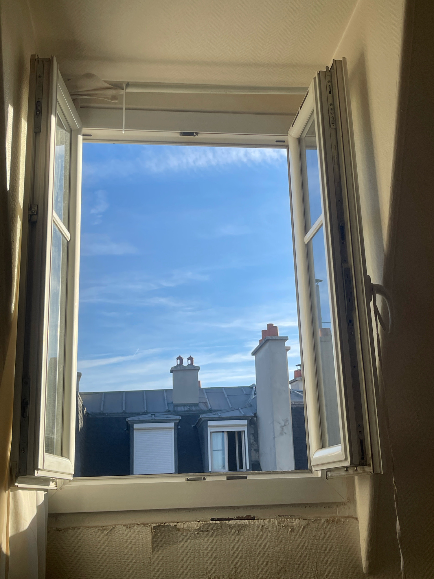 The image shows an open window in Paris, France, looking out onto a cityscape. The window has white frames and is partially open, with the left pane angled slightly inward and the right pane opened wider. Sunlight streams in from the left, casting shadows on the interior wall. Outside, the sky is a clear blue with wispy white clouds. Below the sky are the roofs of buildings, characterized by dark grey shingles. Several chimneys with red-capped tops are visible. One building in the middle has a white shuttered window. The interior sill of the window is visible at the bottom of the frame, with textured wallpaper above it. The scene depicts a typical Parisian architectural view from an apartment window.