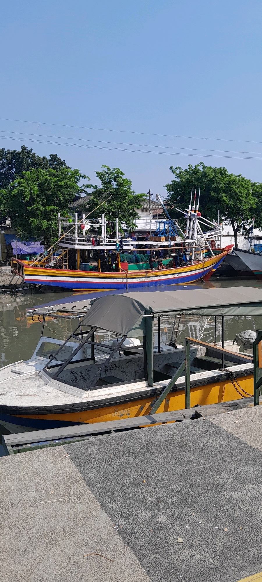 The image shows a harbor in Pasuruan, Indonesia, with several boats docked. In the foreground, a small yellow and white boat with a grey canopy is moored. Behind it, a larger, brightly colored fishing boat with yellow, blue, red, and green stripes is visible. The fishing boat has several people on deck and appears to be loaded with nets and other fishing equipment. Trees and buildings line the shore in the background, and the sky is clear and blue. The overall scene depicts a typical day at a busy port.
