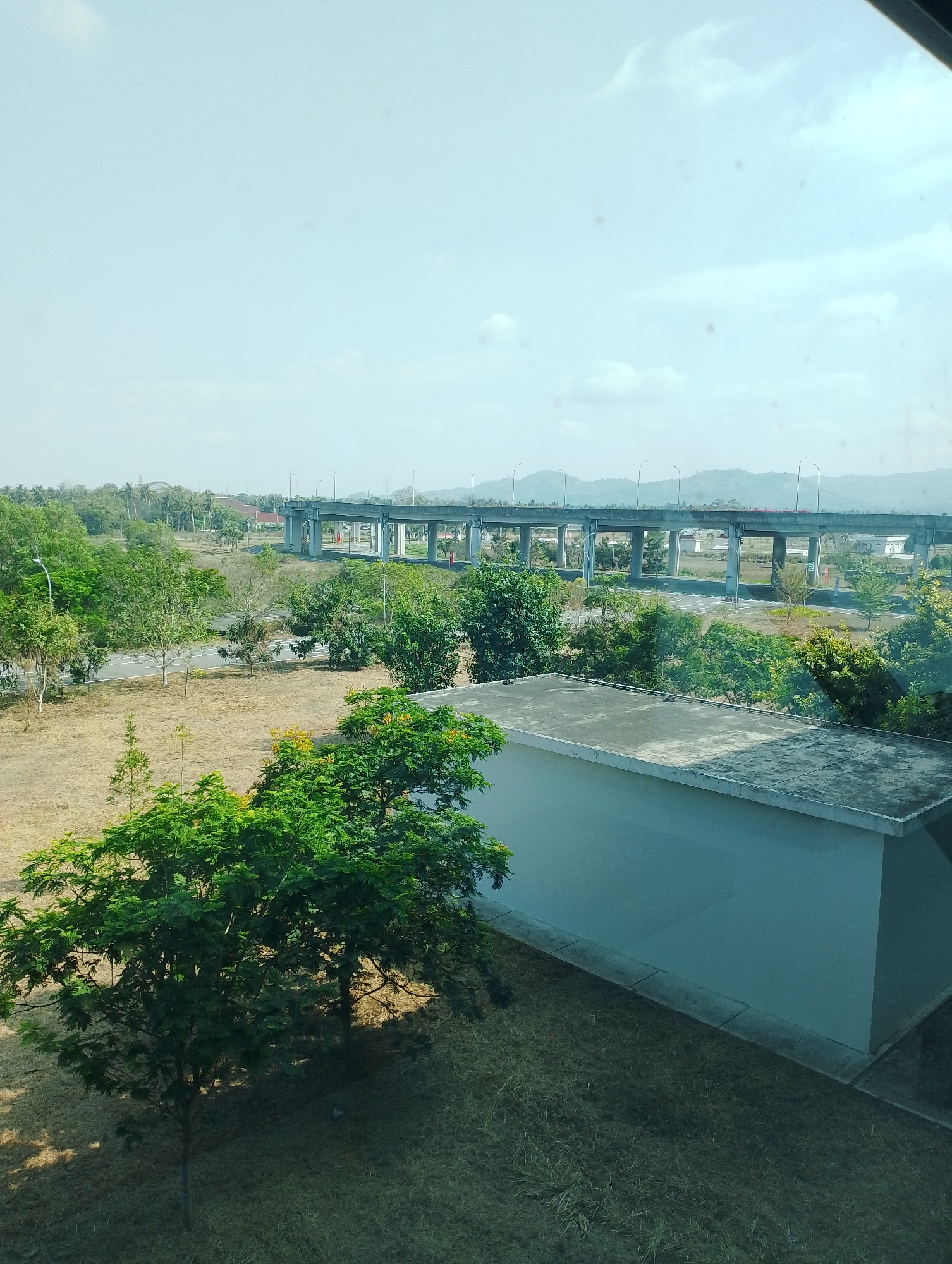 The image captures a panoramic view of an urban or semi-urban landscape under a hazy sky. In the foreground, a low-rise building with a flat, weathered concrete roof and light blue walls occupies the lower right portion of the frame. Adjacent to it, dry, patchy grass covers the ground. To the left of the building, a lush green tree with dense foliage and some yellow flowers stands prominently, casting shadows on the dry grass.
Further into the midground, a paved road with white markings runs horizontally, bordered by more dry grass and scattered trees. A street lamp is visible on the left side of the road. Beyond the road, a substantial elevated highway structure dominates the scene, supported by multiple concrete pillars and featuring a continuous deck. This highway appears to be a significant piece of infrastructure, traversing the landscape and disappearing into the distance.
The background reveals a verdant expanse of trees, including palm trees, suggesting a tropical or subtropical environment. In the far distance, a range of mountains forms a hazy, blue silhouette against the sky, indicating a natural geographical feature. The overall impression is one of a developed area situated within a natural setting, possibly in the vicinity of Palihan, Indonesia, given the vegetation and the architectural style of the highway. The lighting suggests it is daytime, with the sun present but diffused by atmospheric conditions. There are no people or animals visible in the image.