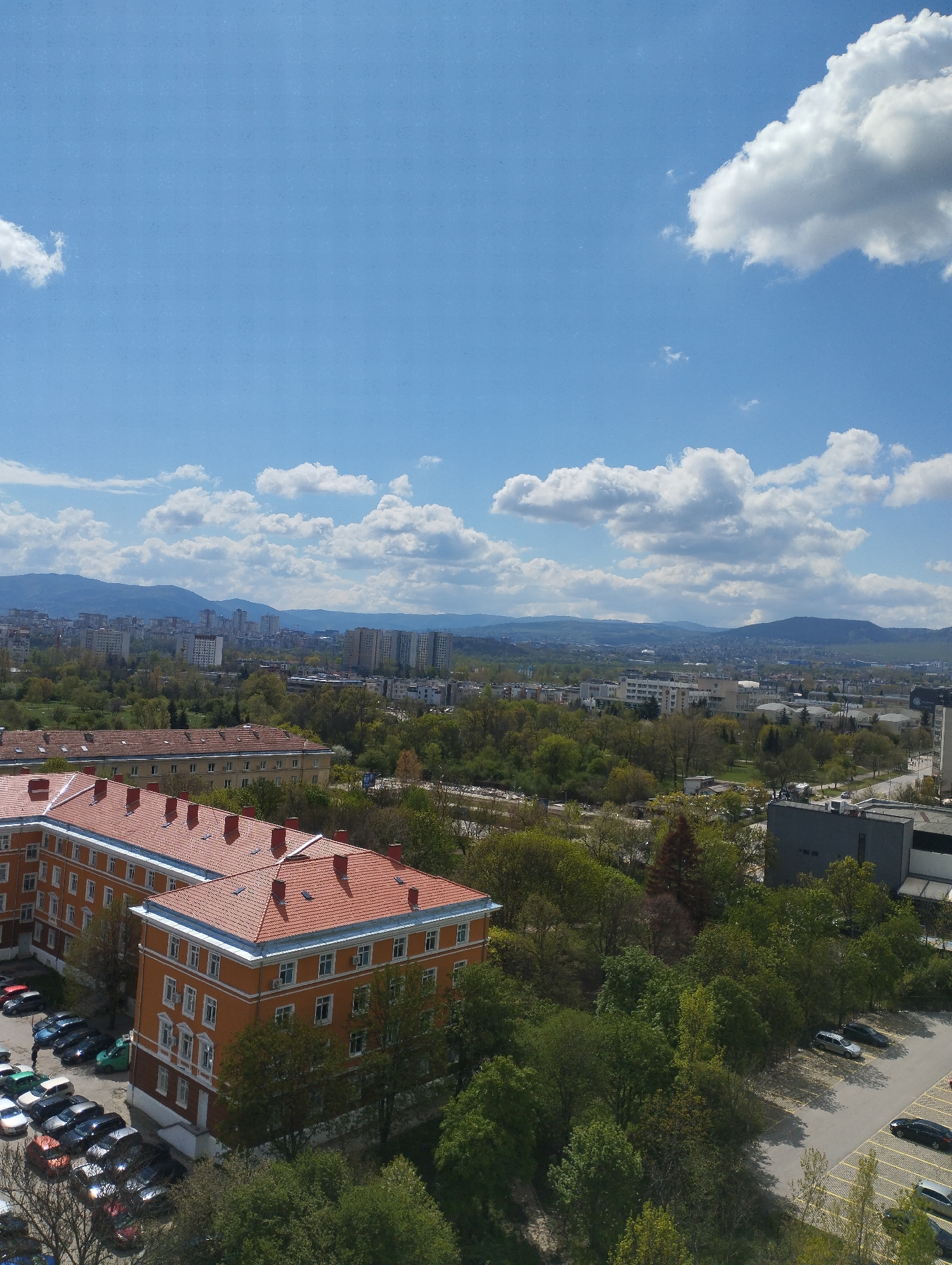 This is a high-angle panoramic photograph of a city or town on a clear, sunny day. The scene is dominated by a bright blue sky dotted with scattered white cumulus clouds.
In the foreground and left mid-ground, a large, multi-winged building stands out with its distinctive orange-red tiled roofs and light-colored (possibly yellow or cream) facades. This building features numerous windows and several roof vents or small chimneys, suggesting an older architectural style. Adjacent to this structure, multiple parking lots are visible, filled with parked cars of various models and colors.
The mid-ground and background reveal an expansive urban area characterized by a mix of residential and commercial buildings, predominantly multi-story apartment blocks. These structures are interspersed with abundant green spaces, including numerous trees with full foliage, indicating a spring or summer season. To the right, a dark, modern building with a flat roof is visible.
A prominent range of mountains forms the distant horizon, framing the cityscape. The time of day is clearly midday or early afternoon, given the bright, even illumination and distinct shadows. The weather appears pleasant, with clear skies and good visibility. No specific activities or people are discernible, and there is no visible text. The overall impression is of a calm, well-established city nestled within a natural, mountainous setting.