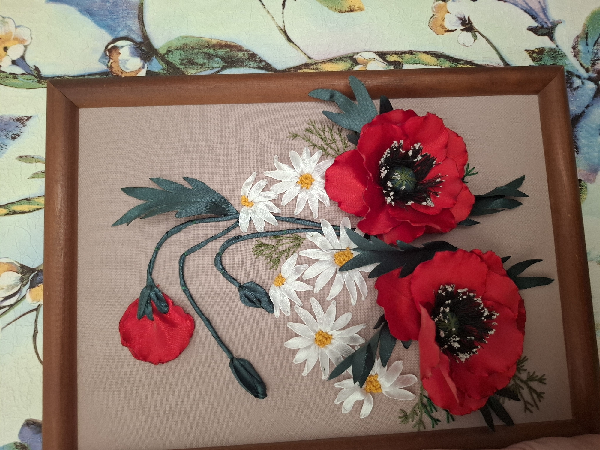 The image depicts a framed textile art piece. The artwork features red poppies, white daisies, green leaves, and poppy buds on a beige fabric background. The frame is wooden with a brown finish. The artwork is positioned against a wallpapered wall with a floral pattern. The image was likely taken in an indoor setting, although the location is unspecified. The scene does not involve people.