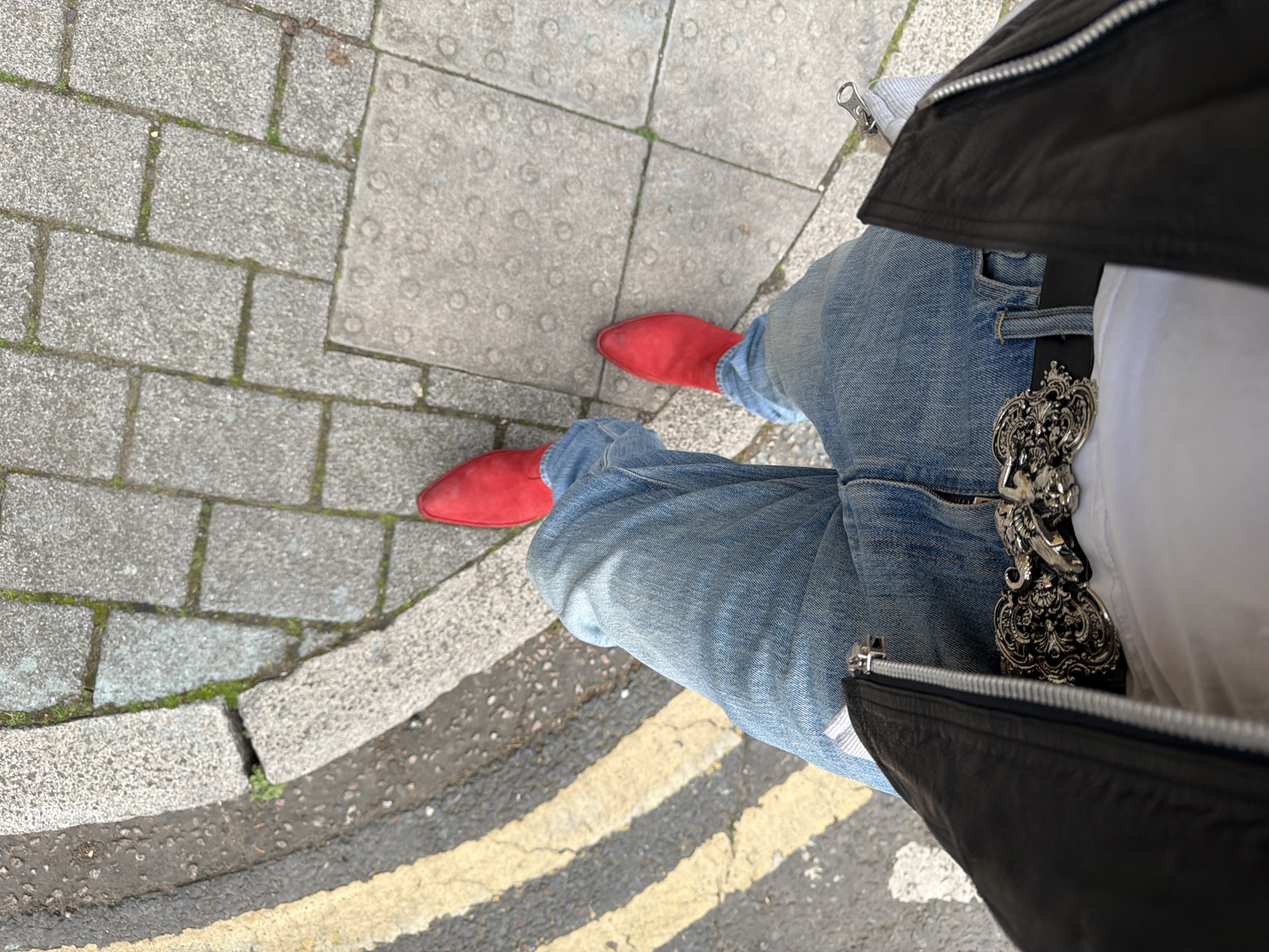 A person stands on a London street. They are wearing red suede shoes, blue jeans, a white t-shirt, a black leather jacket, and a large ornate silver belt buckle. The street has gray paving stones and a double yellow line.