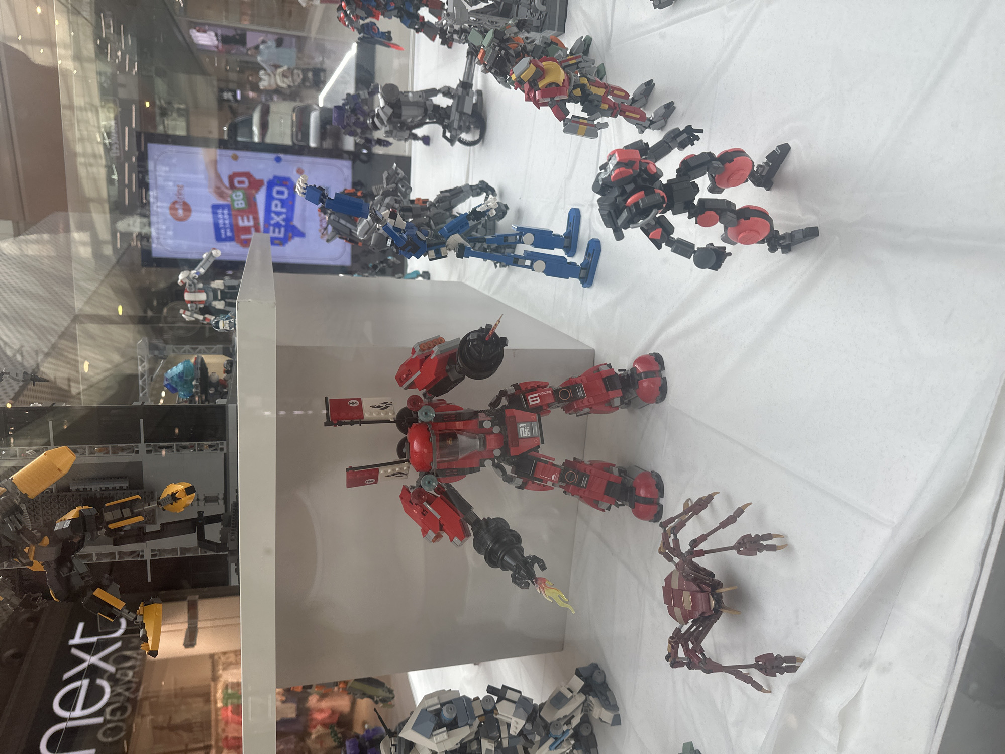 The image displays a collection of intricate LEGO mechs and robots arranged in a storefront window, likely in a shopping mall. A prominent red and black mech stands in the foreground, equipped with a flame accessory protruding from its arm. To its left, a white and blue mech is positioned. A multi-legged, dark red robot resembling a spider is also visible in the lower part of the frame. In the background, other LEGO mechs, some blue, grey, and red, are displayed. A digital screen in the background shows the words "BG EXPO" and the logo of "Telenor". The setting suggests an indoor retail environment, possibly a toy store or an exhibition display, with shoppers visible in the distance through the glass. The overall scene is clean and well-lit, highlighting the detailed construction of the LEGO models.
