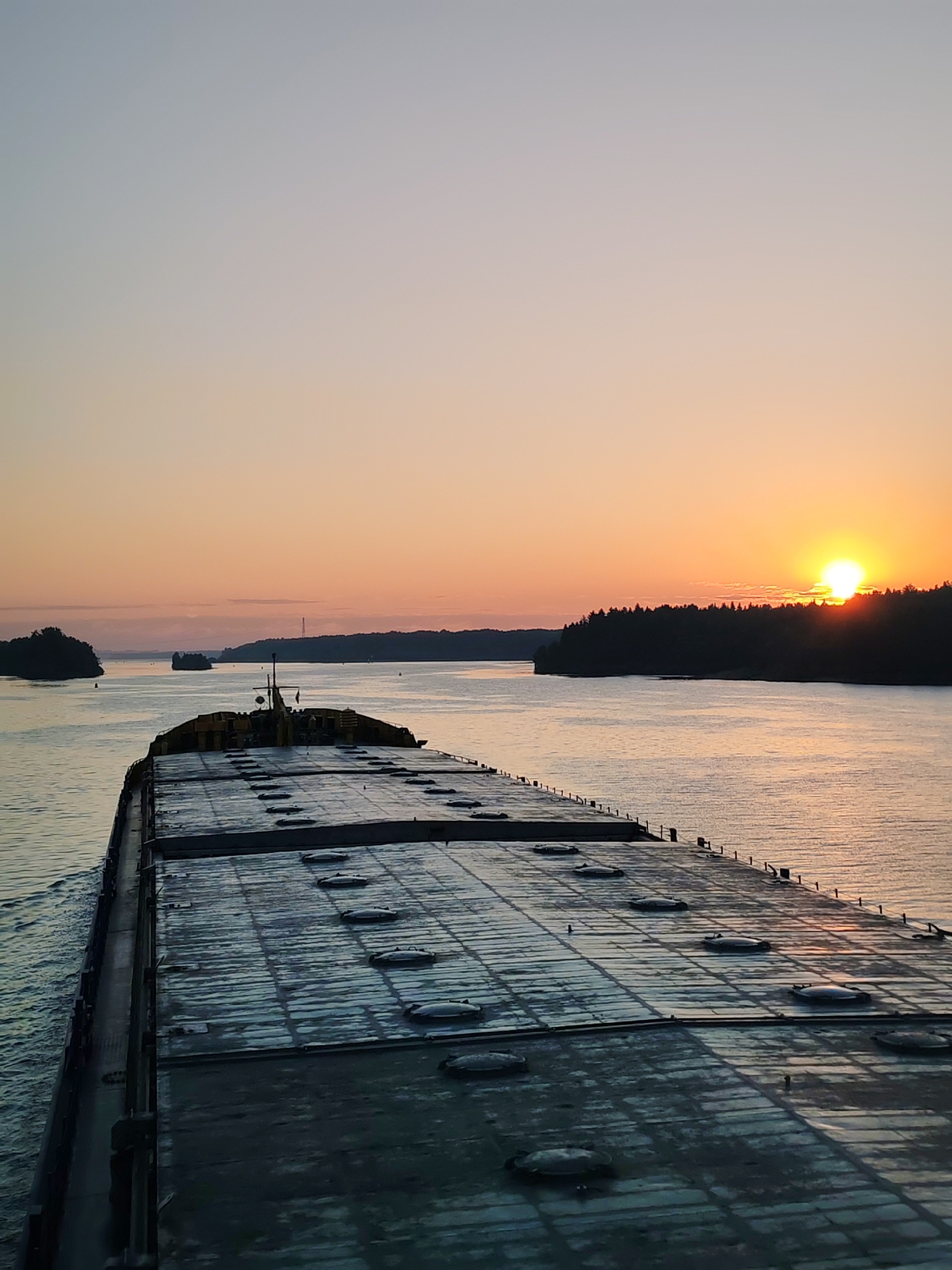 The image depicts a barge traversing a waterway at sunrise. The barge is large, gray, and flat, with a grid pattern of hatch covers across its deck. A yellow structure sits atop the barge towards the center of the frame. The water reflects the warm hues of the rising sun. The river is flanked by land on either side, with dense forests on the right and smaller islands on the left. Another vessel can be seen in the distance. The sky is clear and transitions from light blue to orange near the horizon. The location is unspecified and information about it being in Poteryaevo, Russia, is not provided within the image.